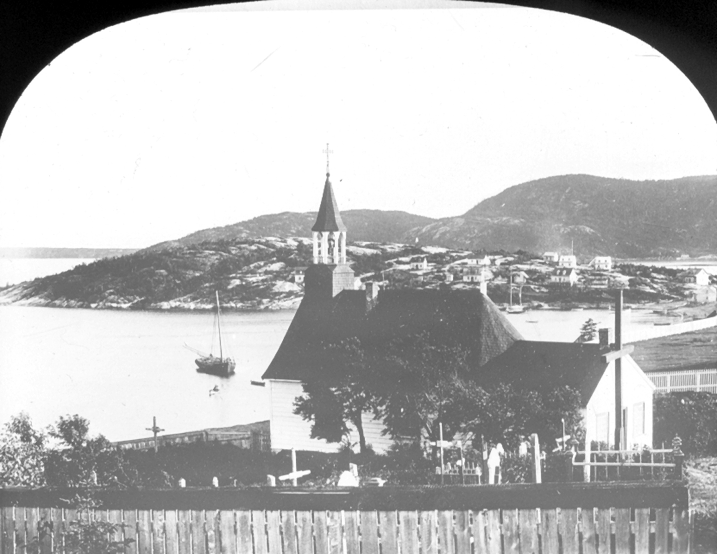 Une chapelle et son cimetière dominent une colline. Quelques maisons se trouvent sur la côte. Un bateau à voile est amarré.