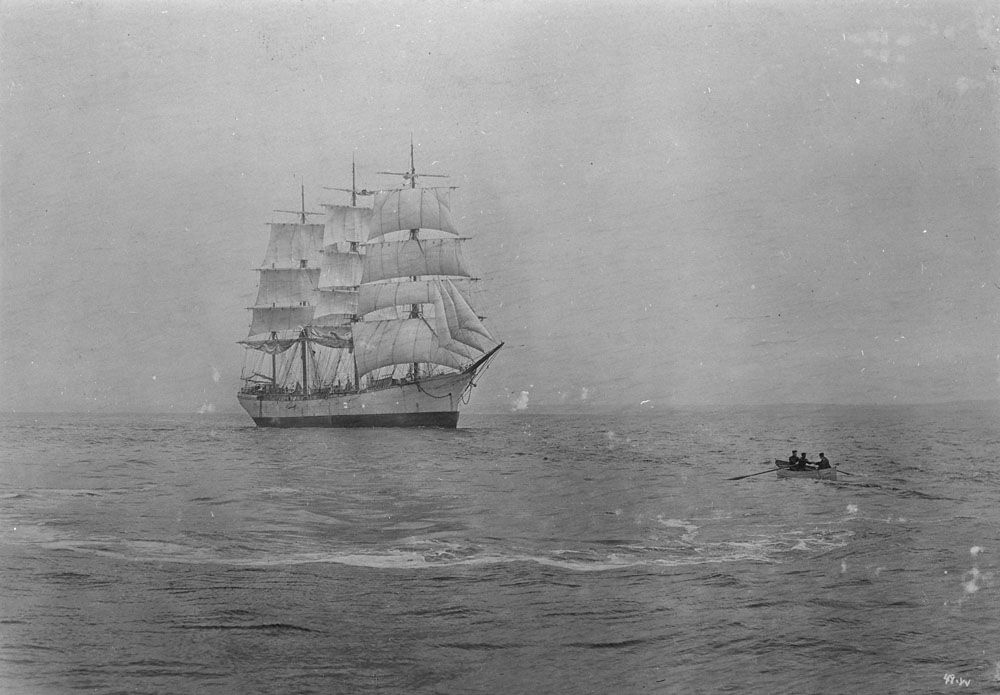 Photographie ancienne en noir et blanc d'une petite embarcation qui se dirige vers un navire à voile