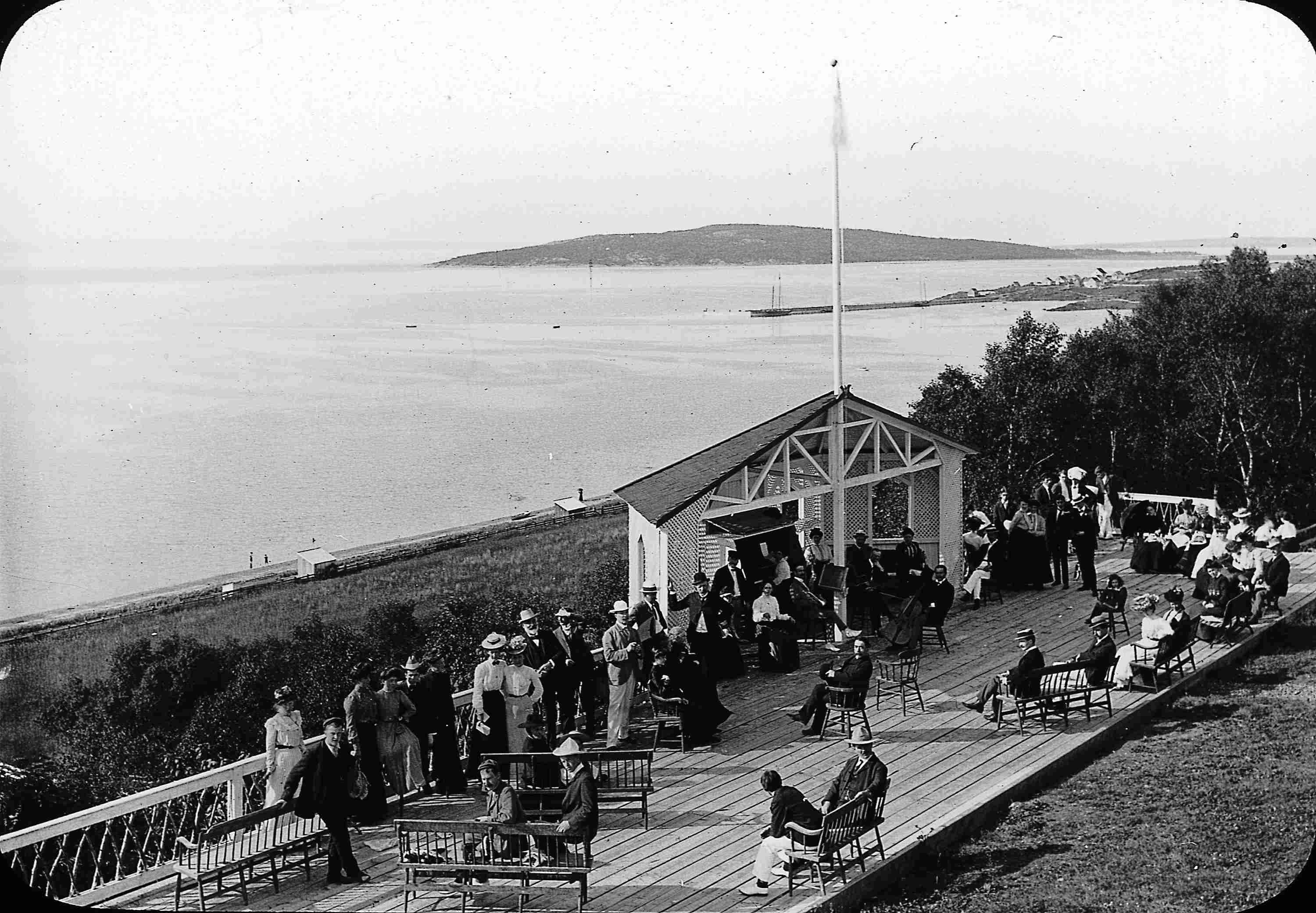 Vacationers conversing on a wood terrace built on a cliff overlooking the water.