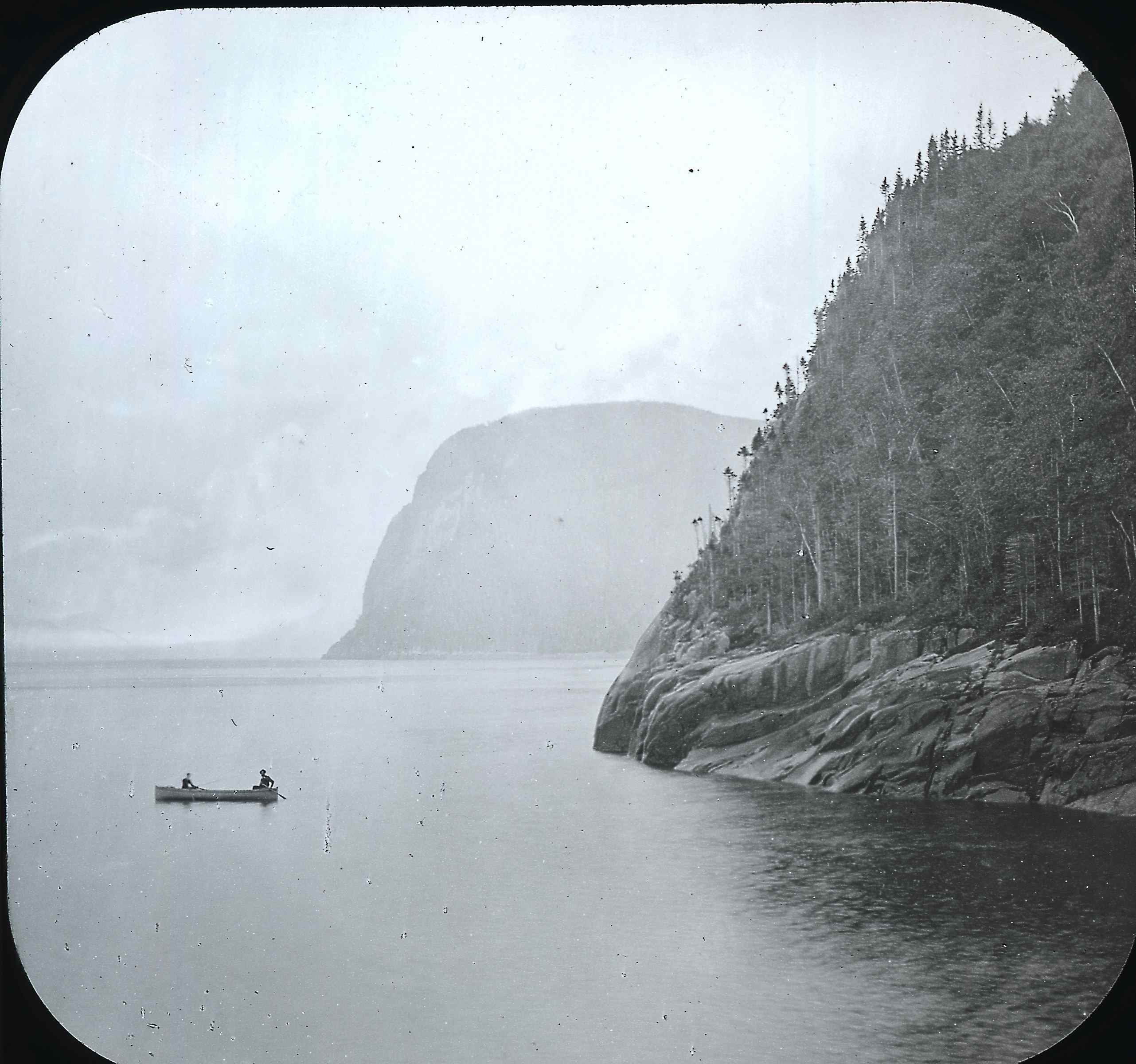 Two canoers fishingat the foot of a cliff that plunges into a very wide river.