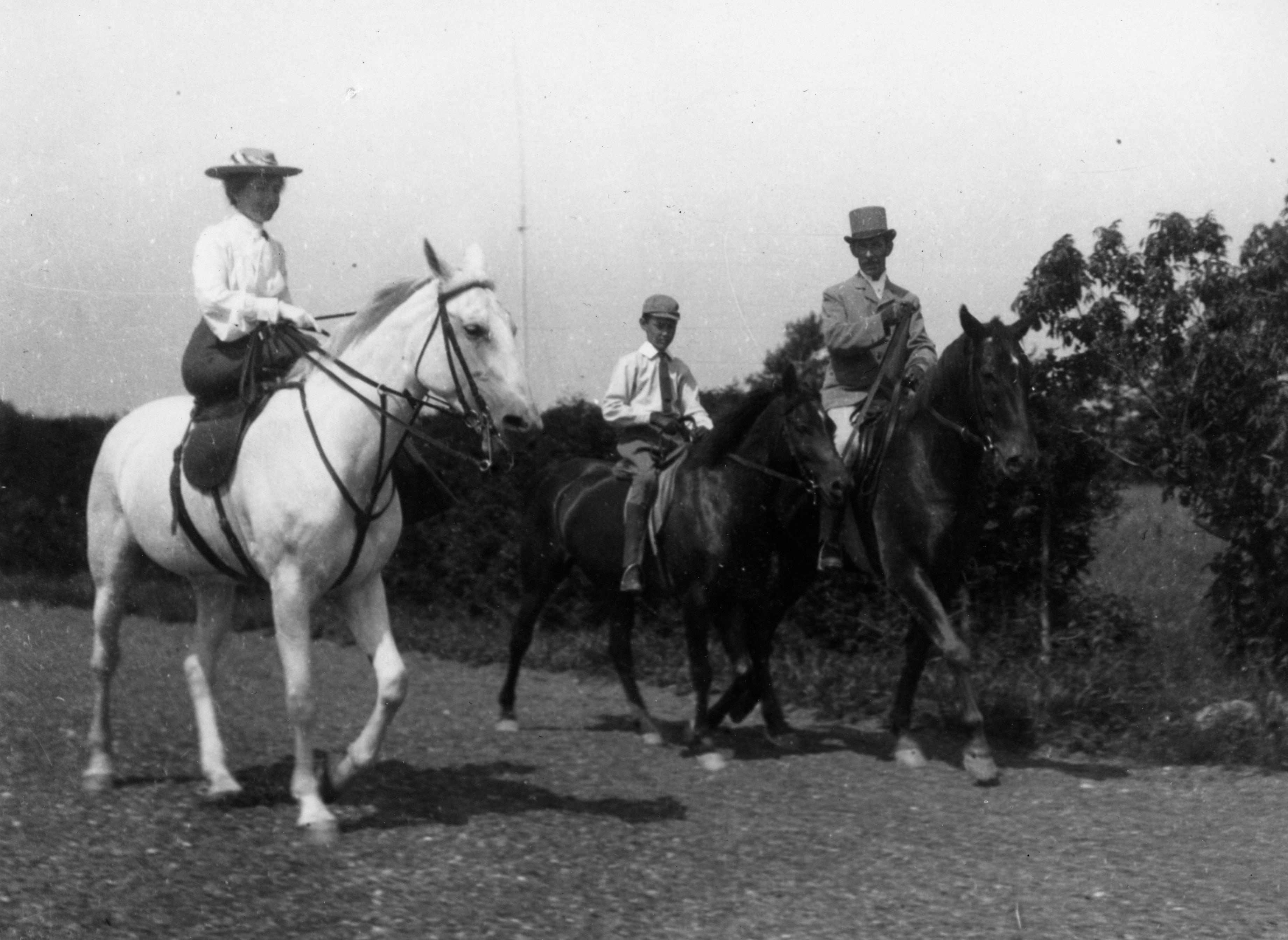 A photograph of three riders on a road: a woman mounted side-saddle, a child, and another elegant horseman.