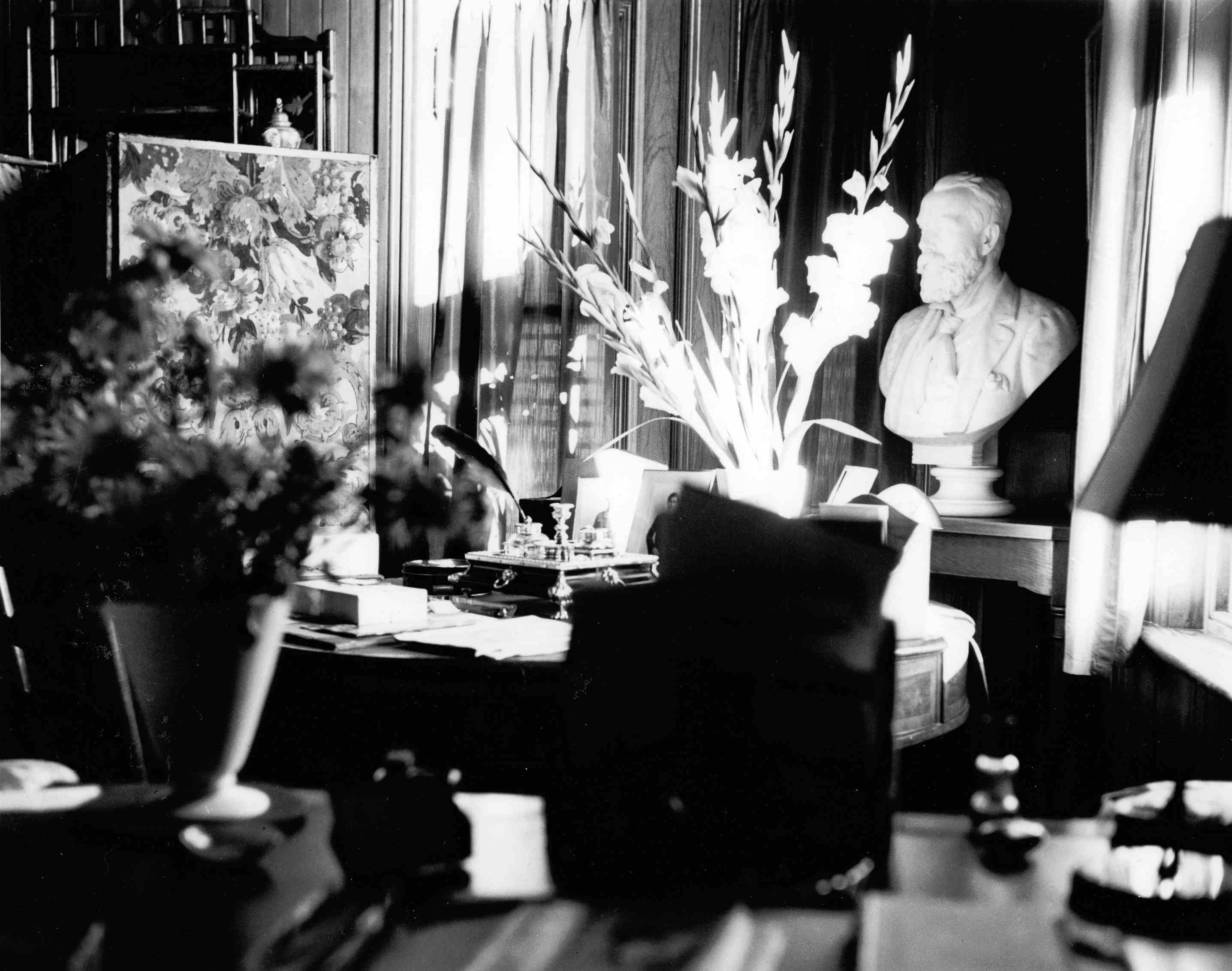 Photographie en noir et blanc d’un bureau à la décoration chargée de bibelots et de fleurs.