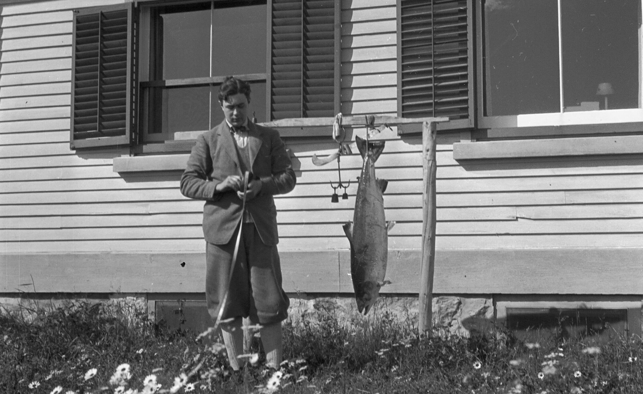 A young man rolls up a measuring tape beside an outdoor scale for weighing fish. A salmon is hanging from the scale.
