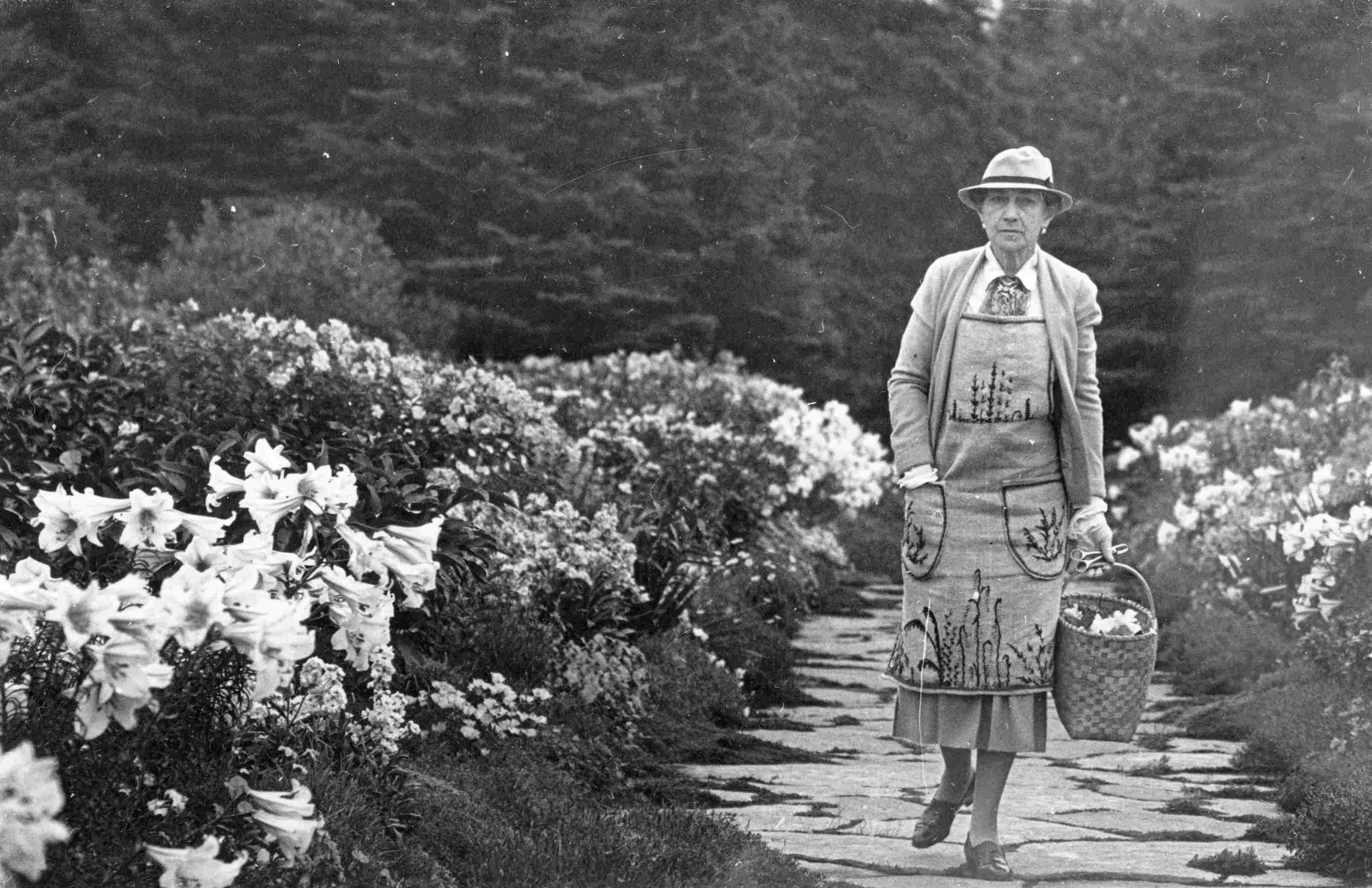 Une femme distinguée, vêtue d’un tablier brodé, marche dans une allée fleurie, un panier à la main.