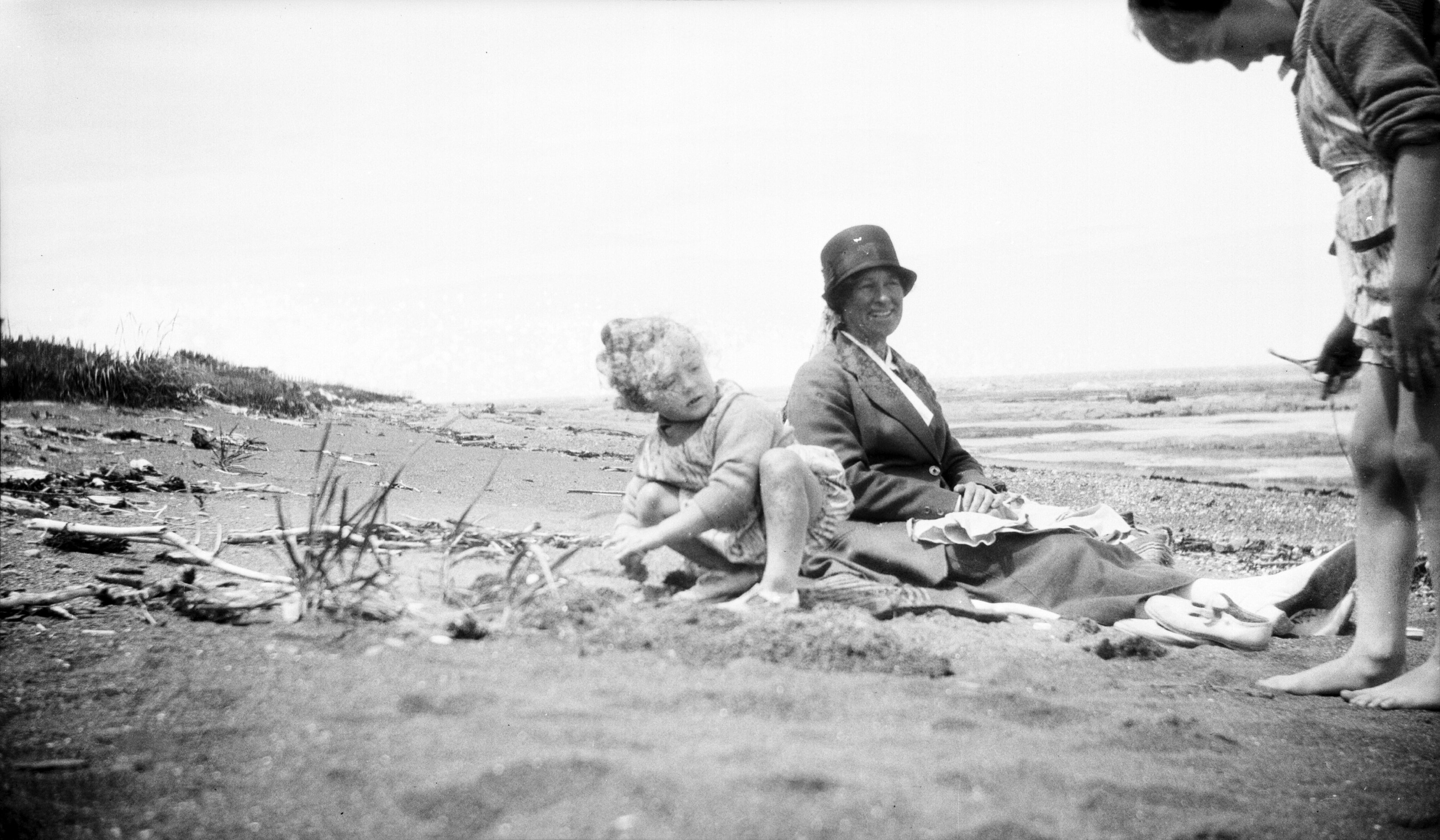 Two children play on the beach at low tide, watched over by a seated woman.