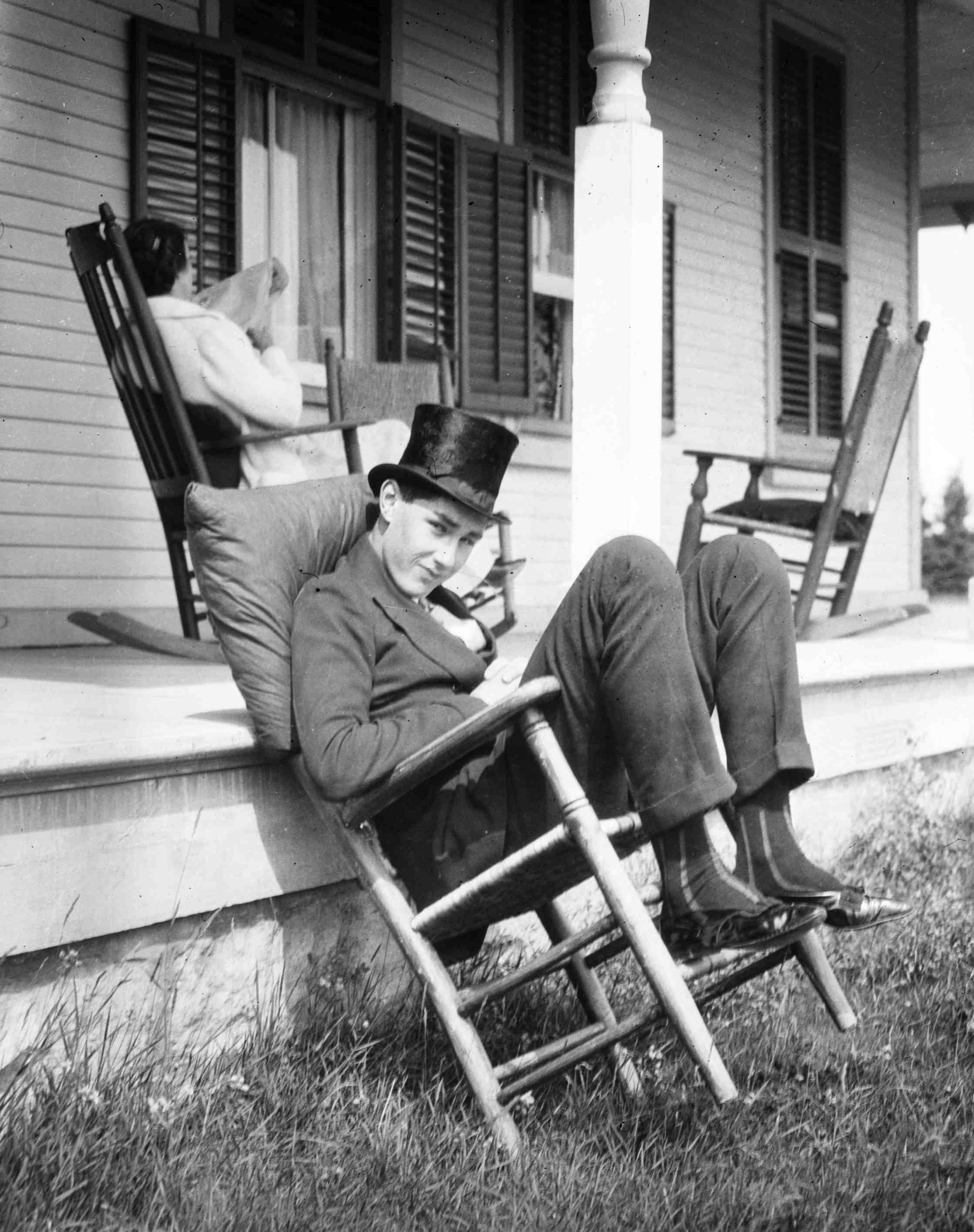 A young man wearing a top hat and sitting nonchalantly on a chair, while a woman reads in the background.