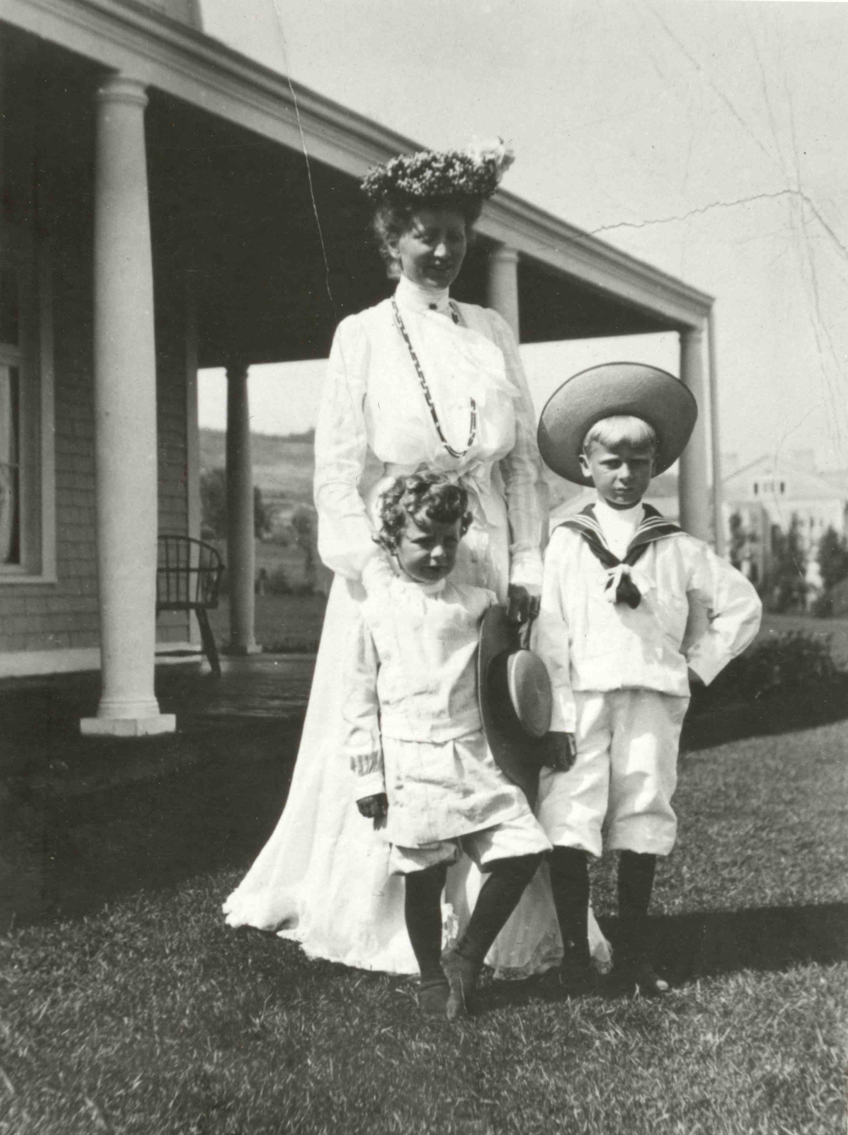 A woman in a very elegant white dress posing with her two children near a house.