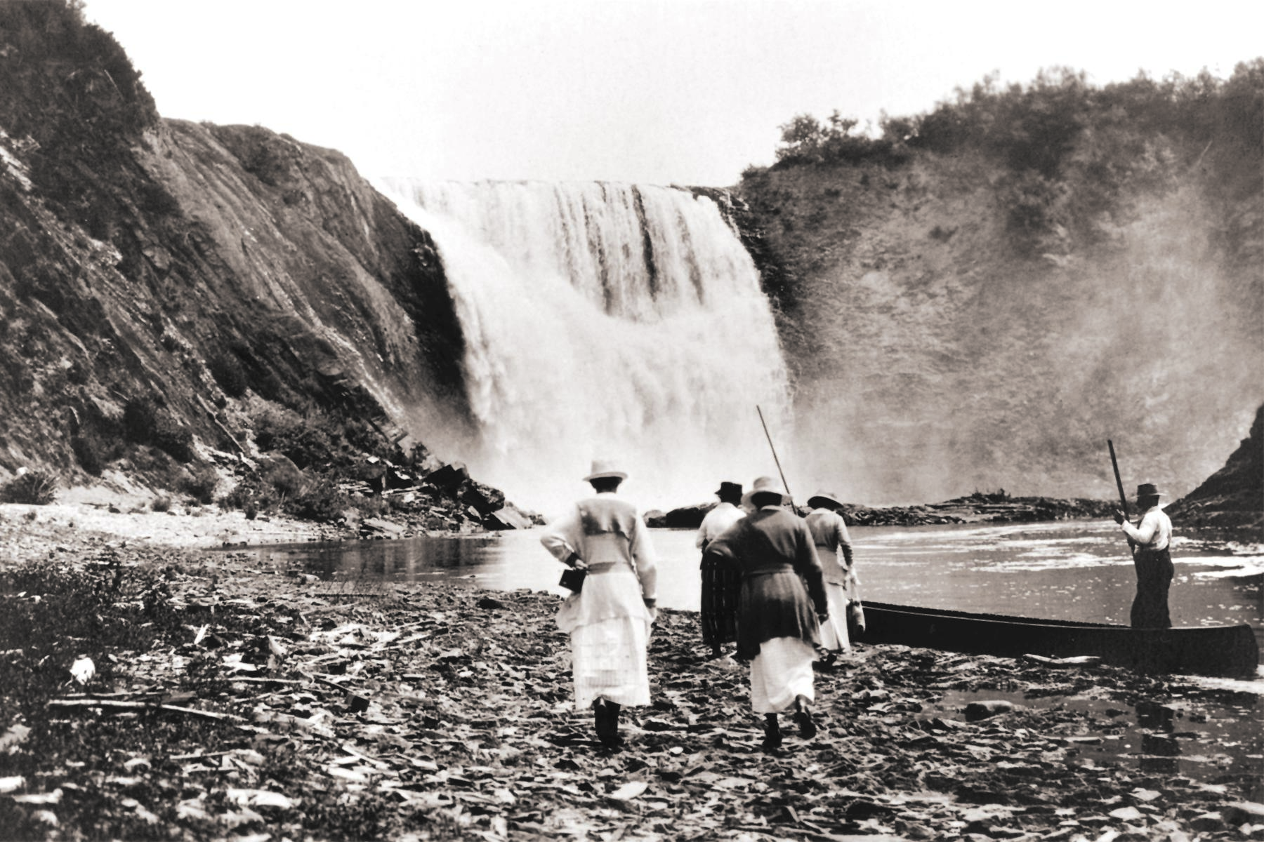 Four women walking at the foot of a waterfall, heading toward a guide standing in a canoe.