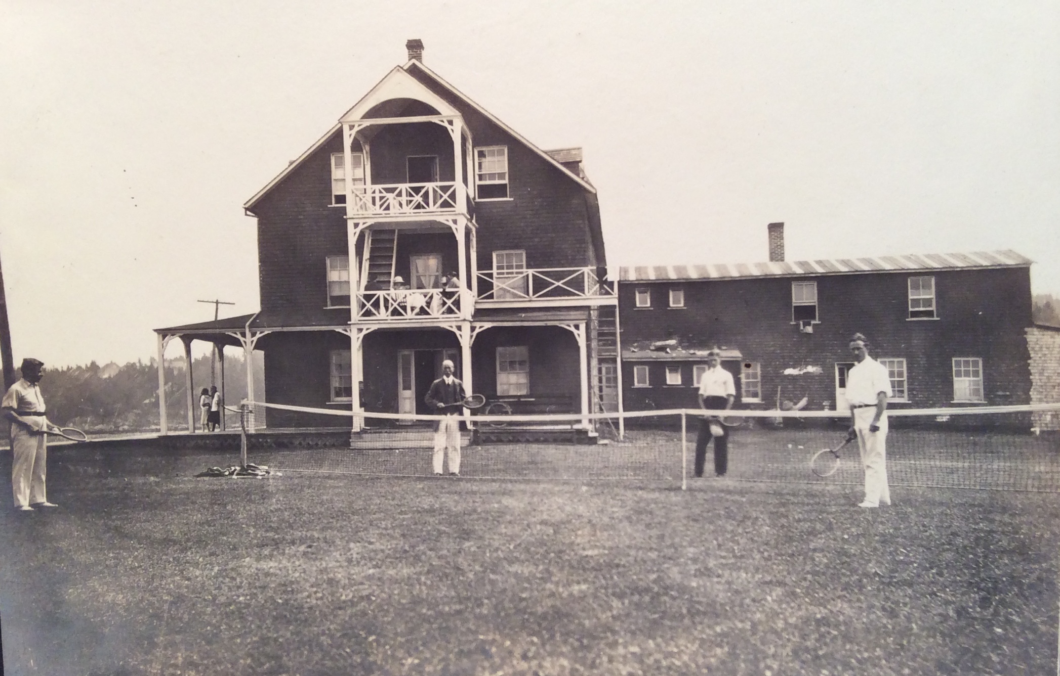 Four men play tennis on the lawn of a hotel on the shore of the St. Lawrence.