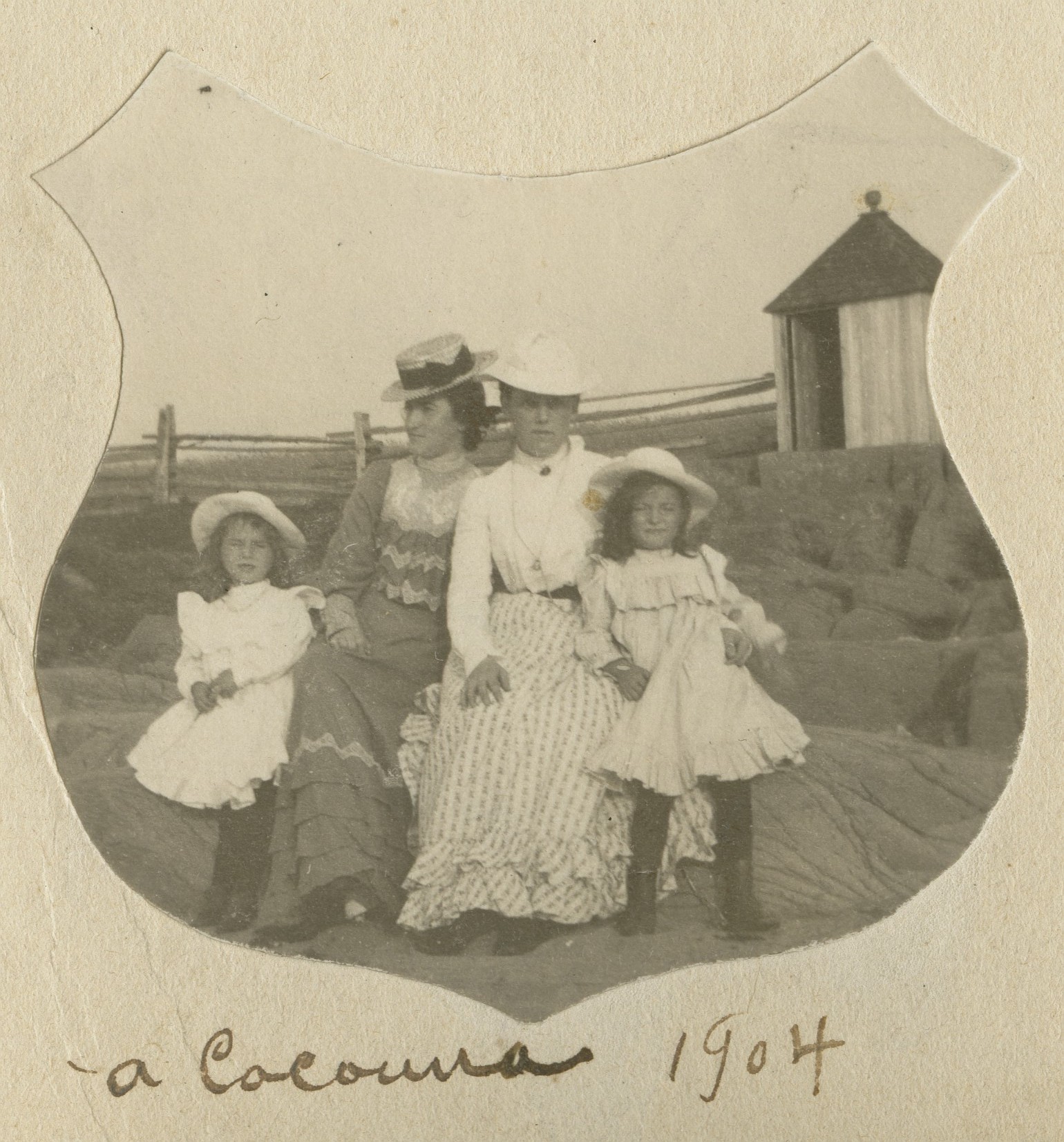 Two young women and two girls pose on rocks by the shore. A changing-cabin is in the background.