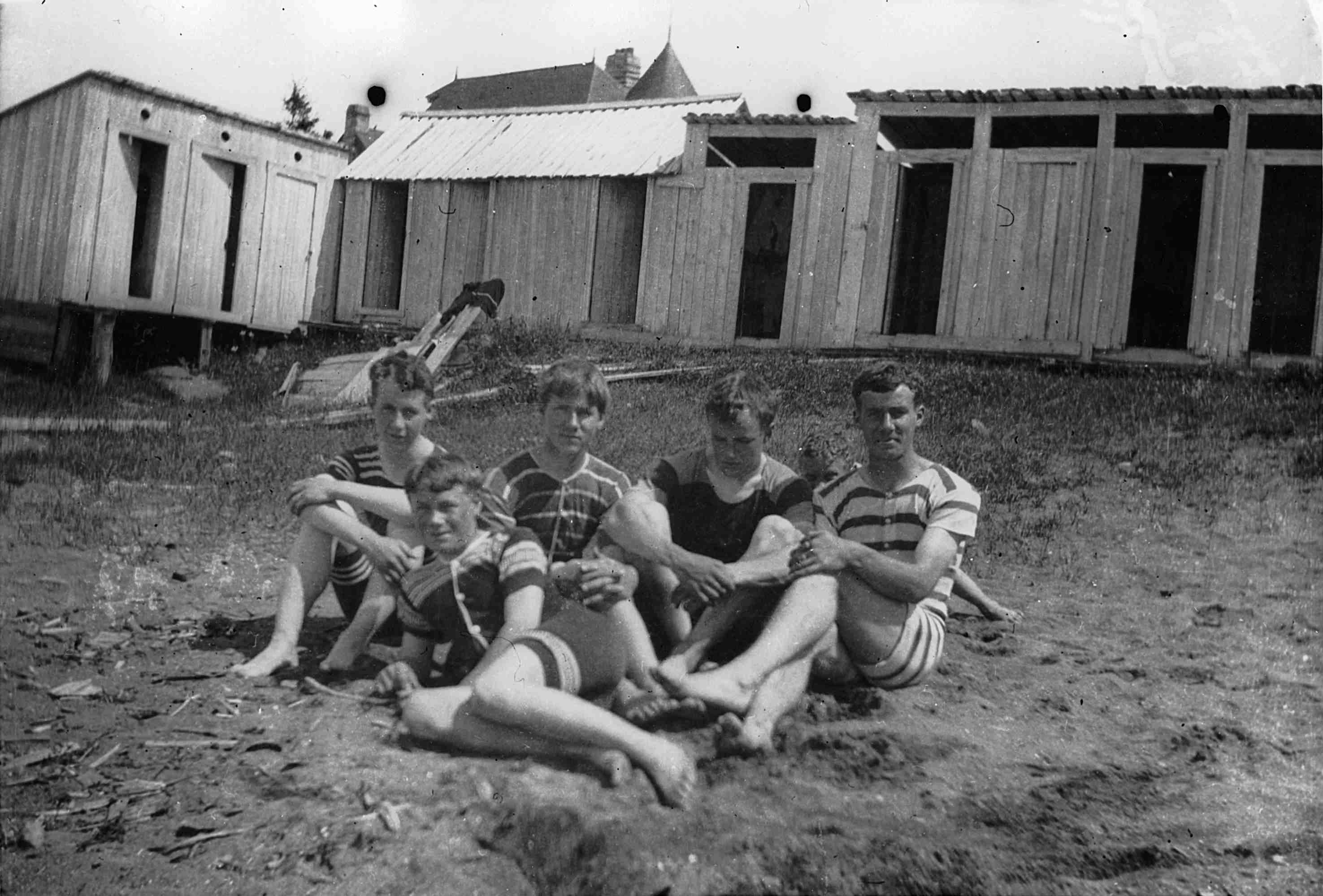 Five adolescents wearing old-fashioned bathing suits and sitting in the sand near a row of bathing cabins.