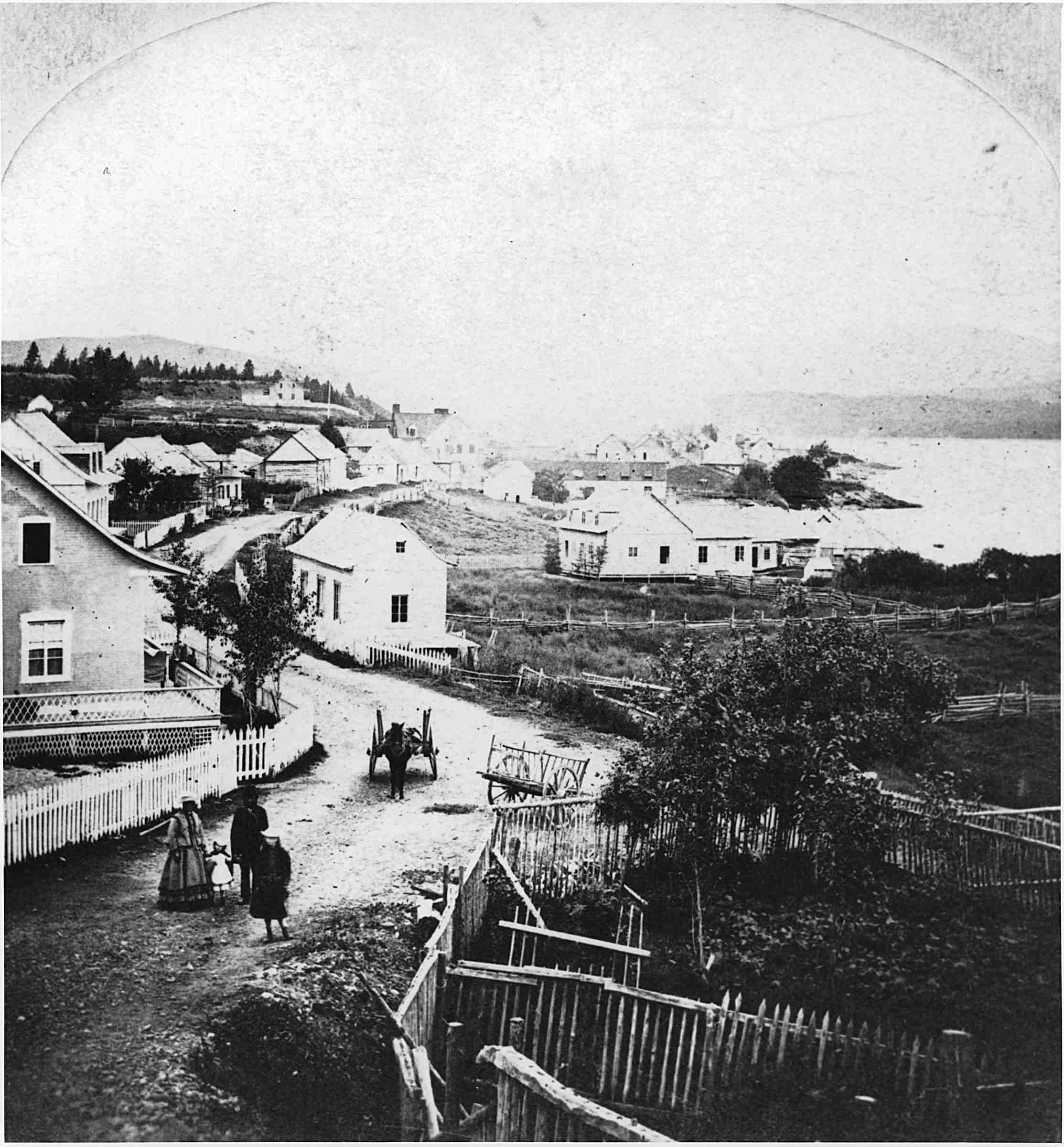 An old photograph of a hilly seaside village. A few people are in the street, near a harnessed horse.