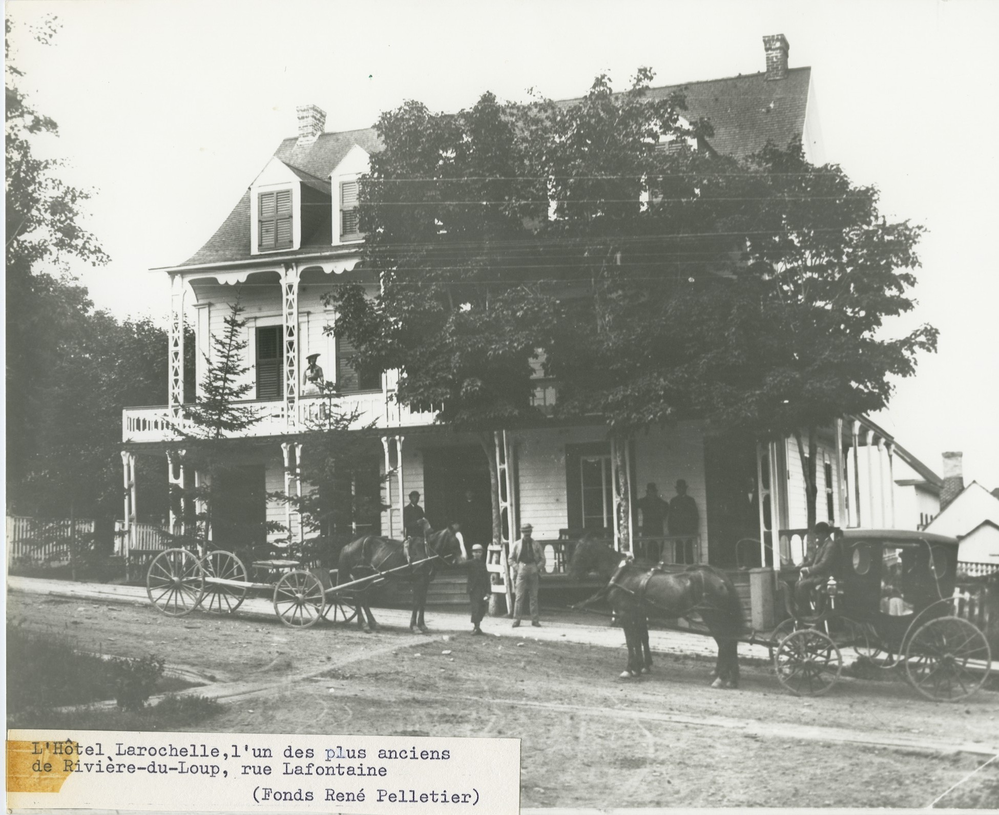Photograph of a small hotel, with two horse-drawn carriages parked in front.
