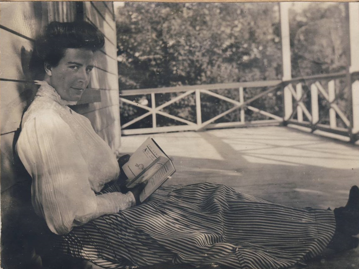 A woman reading, sitting on the floor of a broad porch, her back against the wall of the house.