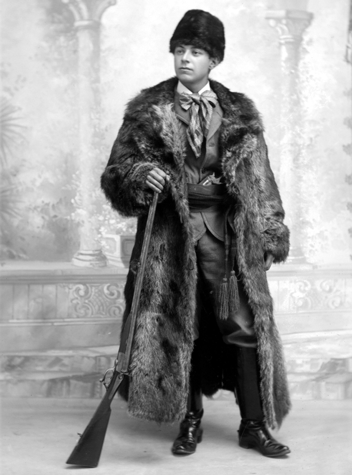 A young man wearing a fur coat and hat, and holding a hunting rifle in his hand.