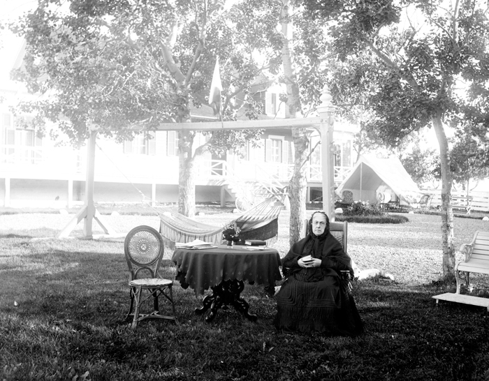 Une vieille femme lit, assise sur une chaise disposée sur le gazon.