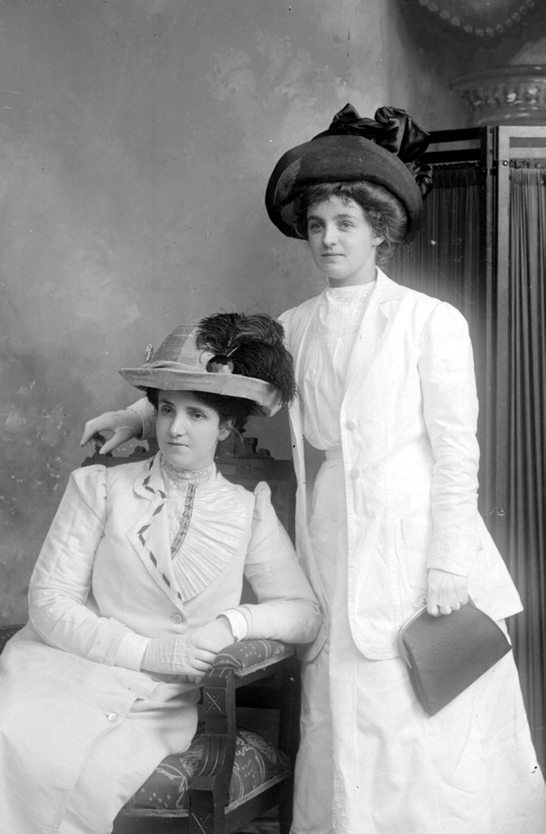 Black and white photograph of two elegant women wearing all white or very pale dresses, including gloves.