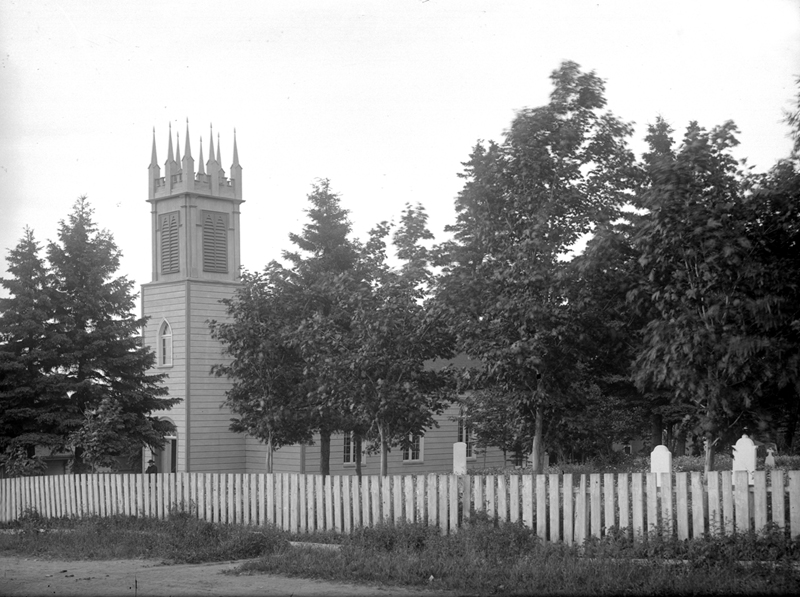 Vue sur une petite église au clocher de style néo-gothique, avec le cimetière attenant. Le terrain est fermé par une clôture.