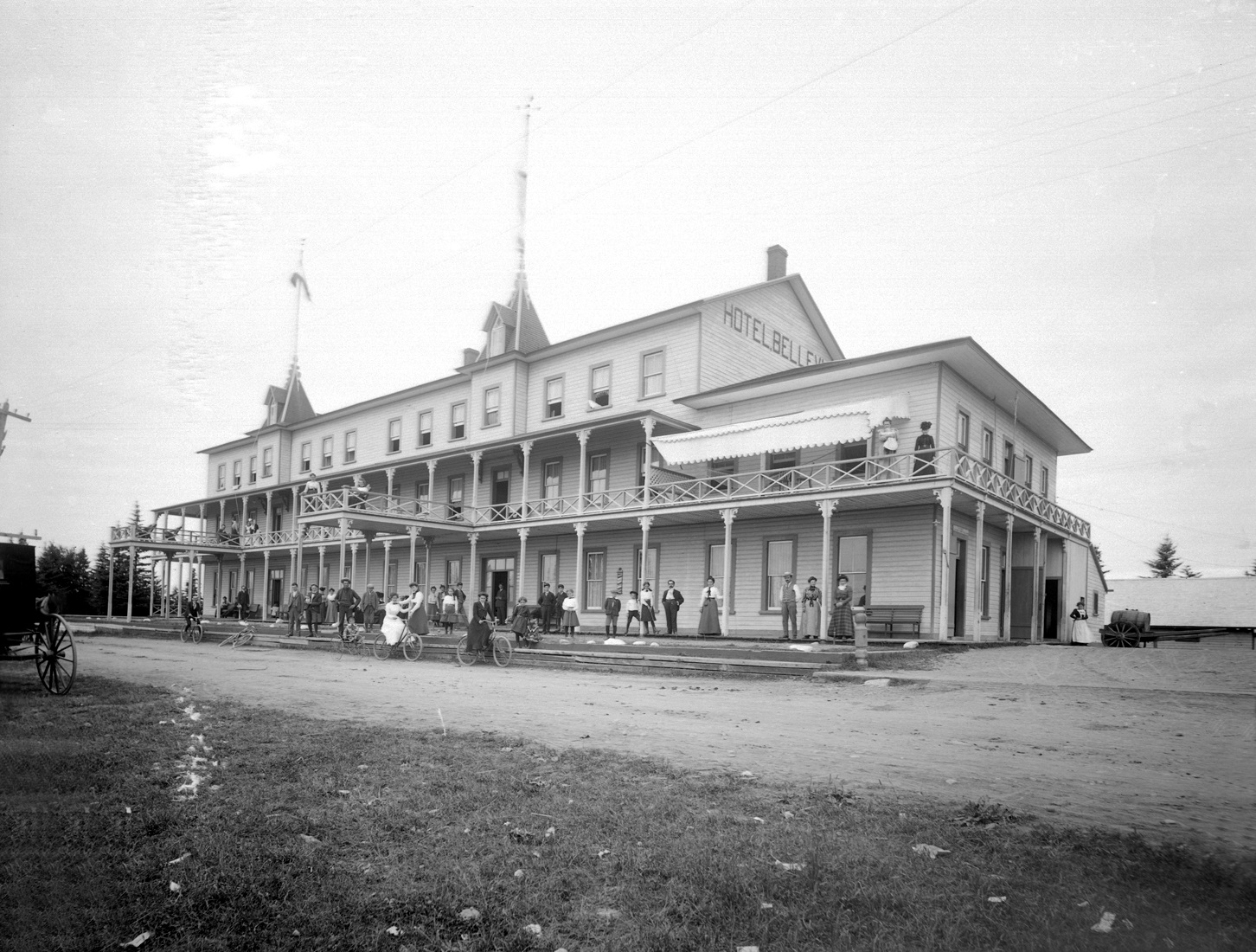 Grand hôtel de bois ceinturé par une grande galerie. Plusieurs touristes, dont des cyclistes, posent sur le perron de l’hôtel.