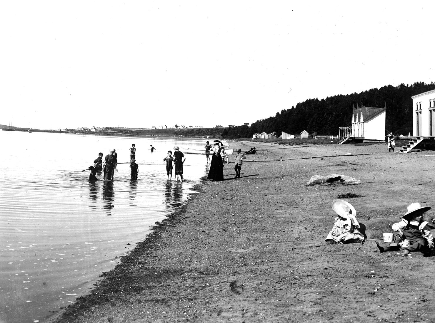 Des femmes et des enfants profitent de la plage. Deux enfants jouent dans le sable. Les baigneurs portent des maillots longs.