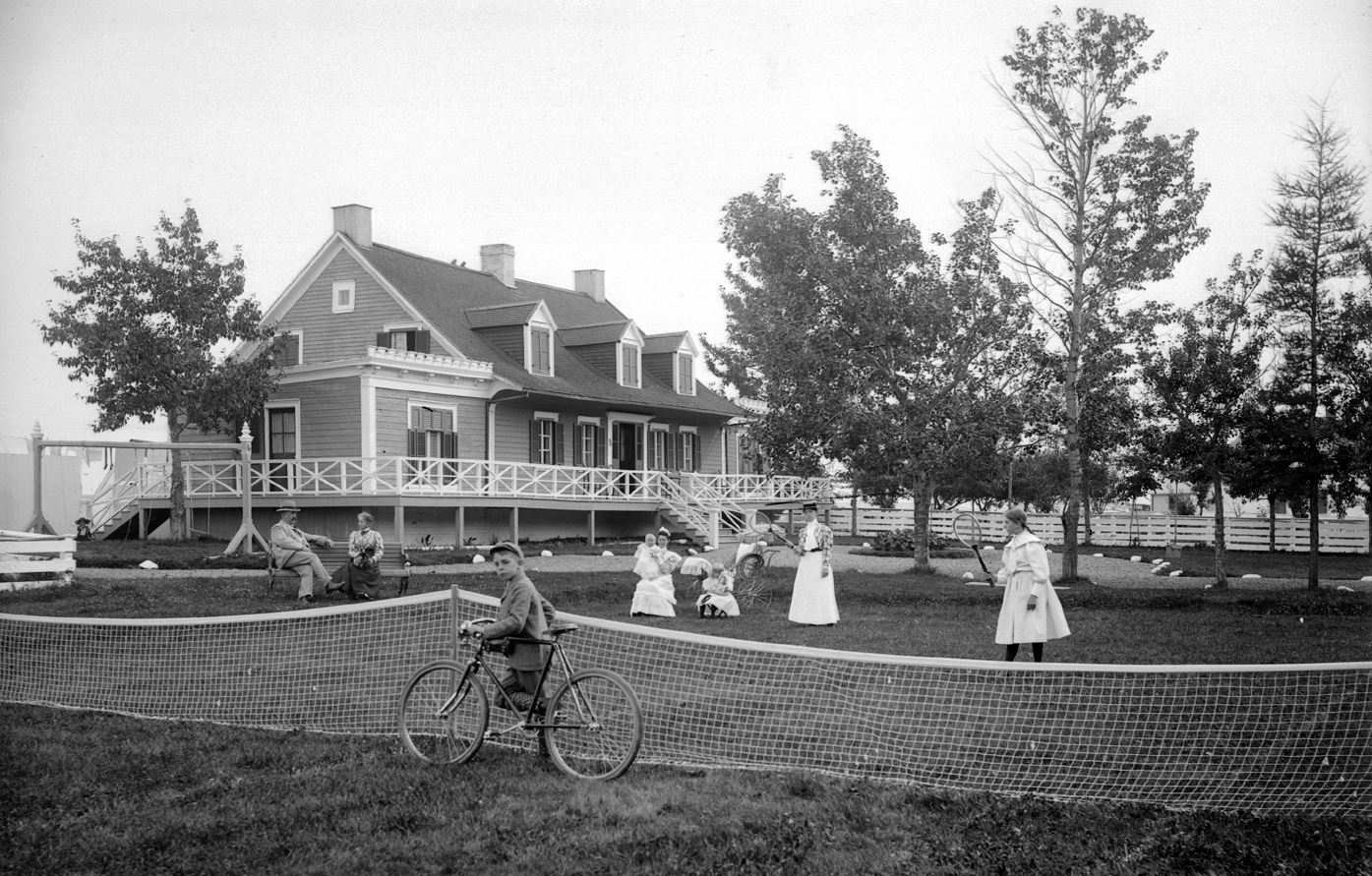 A family enjoying summer in front of a posh house and a child riding a bicycle, while others play tennis or talk.