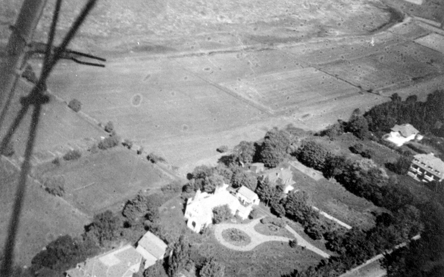An aerial photograph in which we see a wooded estate near large fields.