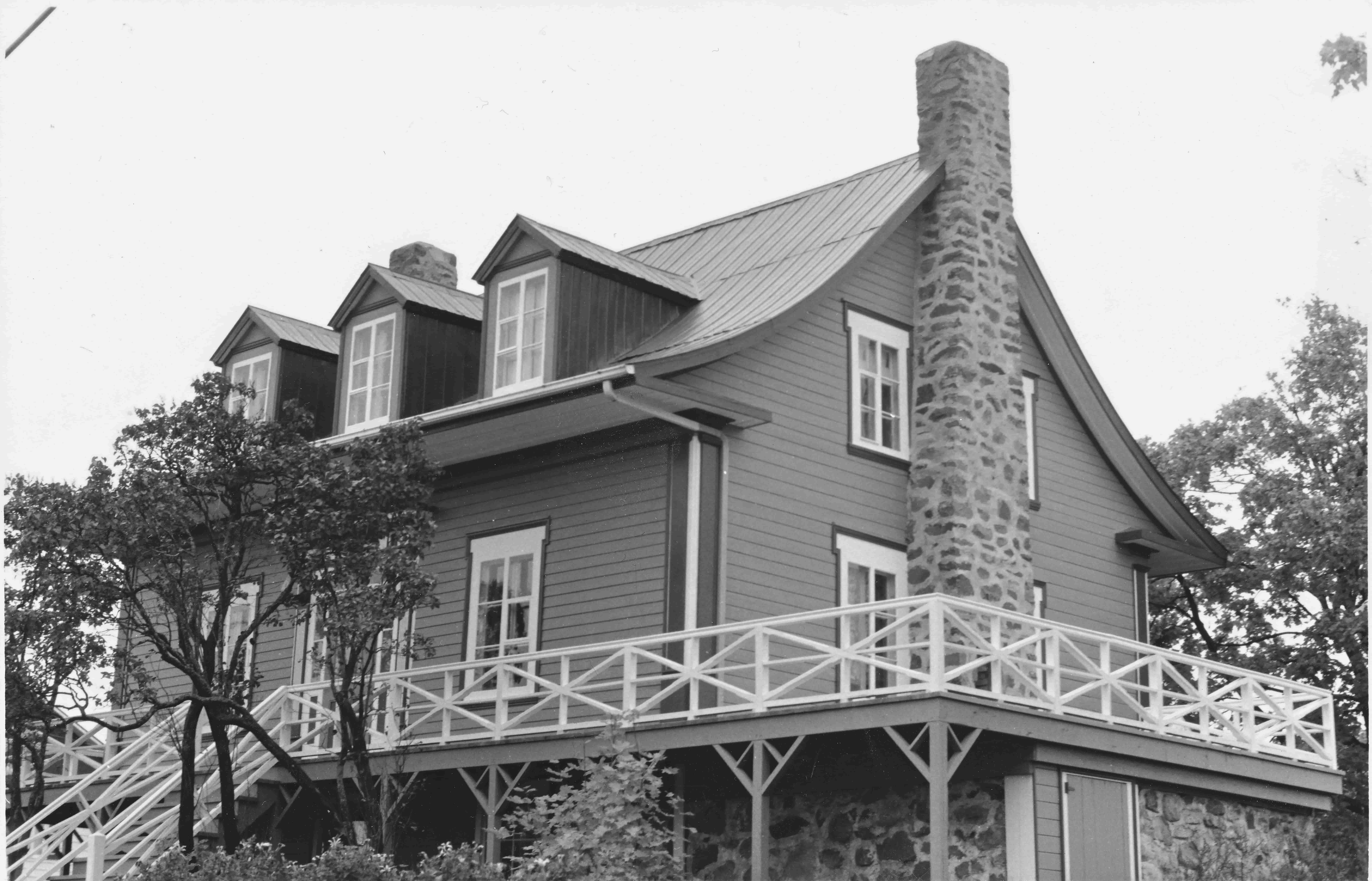 Solide maison québécoise ancienne, avec cheminée de pierre, toit en tôle et murs de planches horizontales en bois.