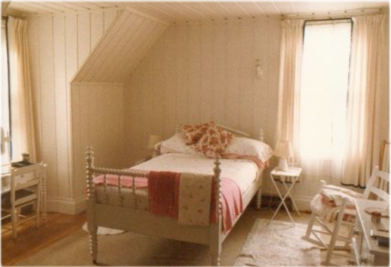 Chambre où se trouvent un lit à une place dont la base est en bois tourné, une commode, une berçante et un petit bureau.