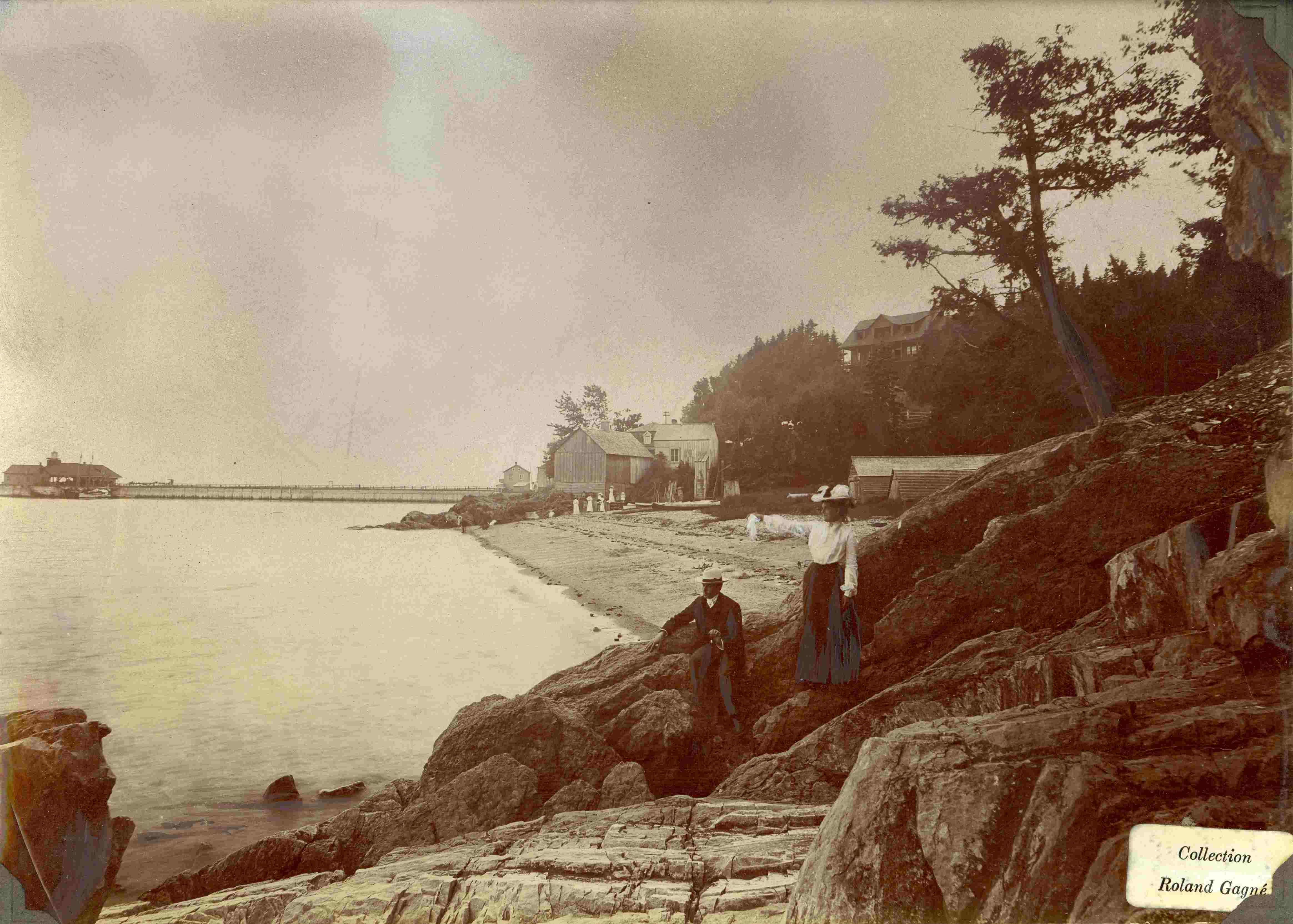 A couple of vacationers have climbed onto the rocks; a beach and a long wharf are in the background.