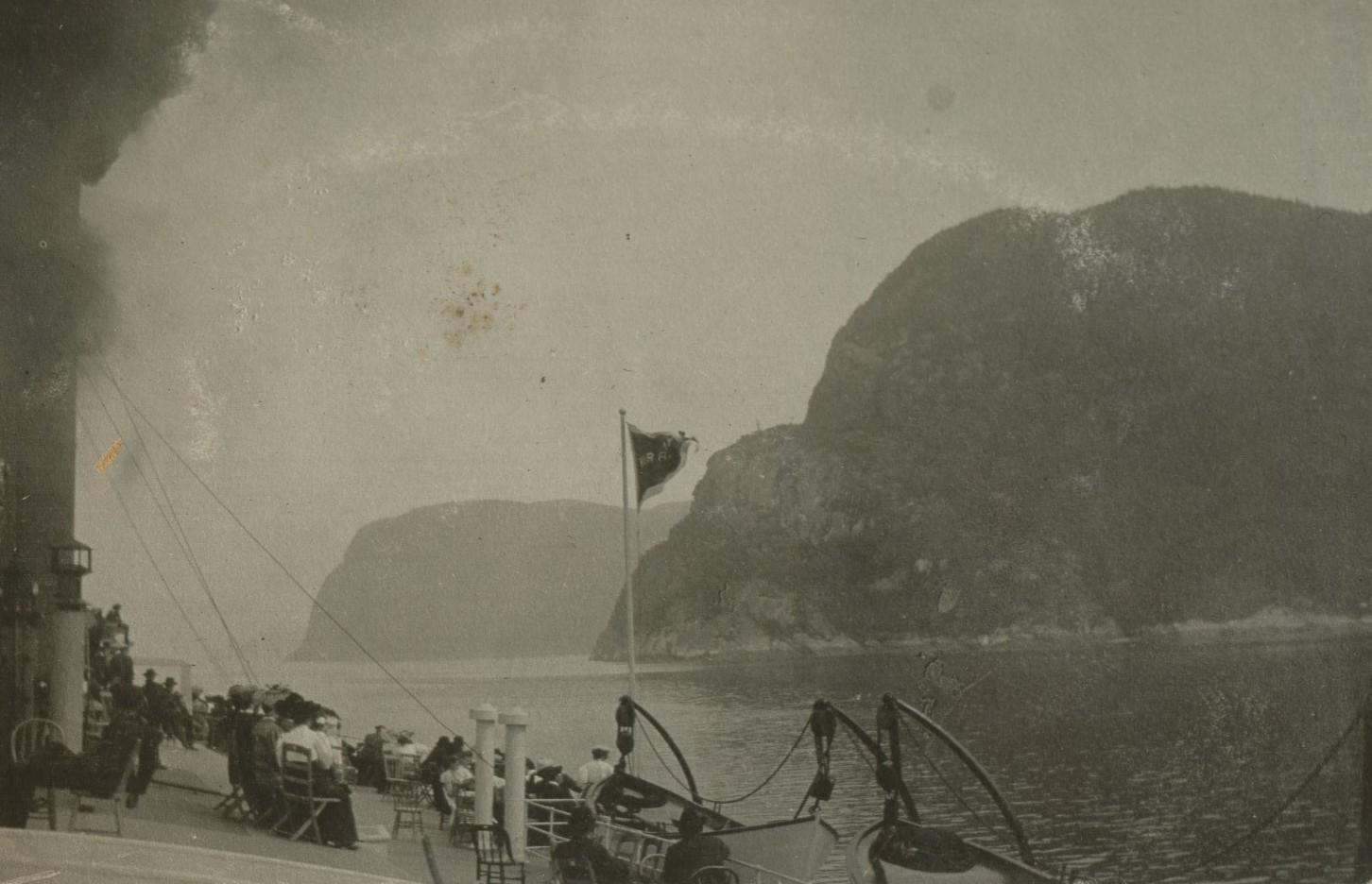 Passengers sitting on the bridge of a steamer, looking at the steep mountains on the shore.