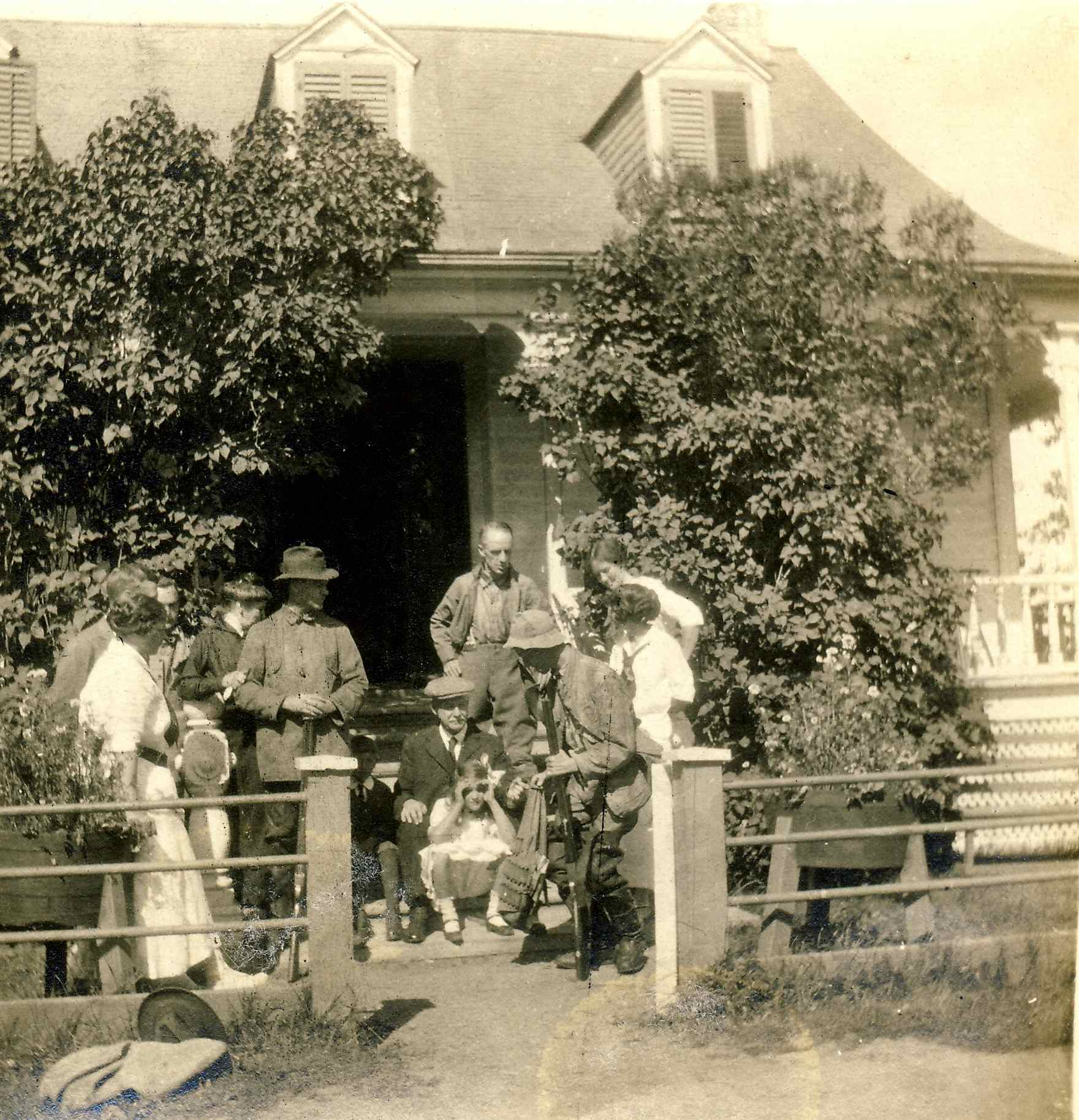 Deux chasseurs sont entourés de quelques personnes devant une maison rurale bordée de lilas.