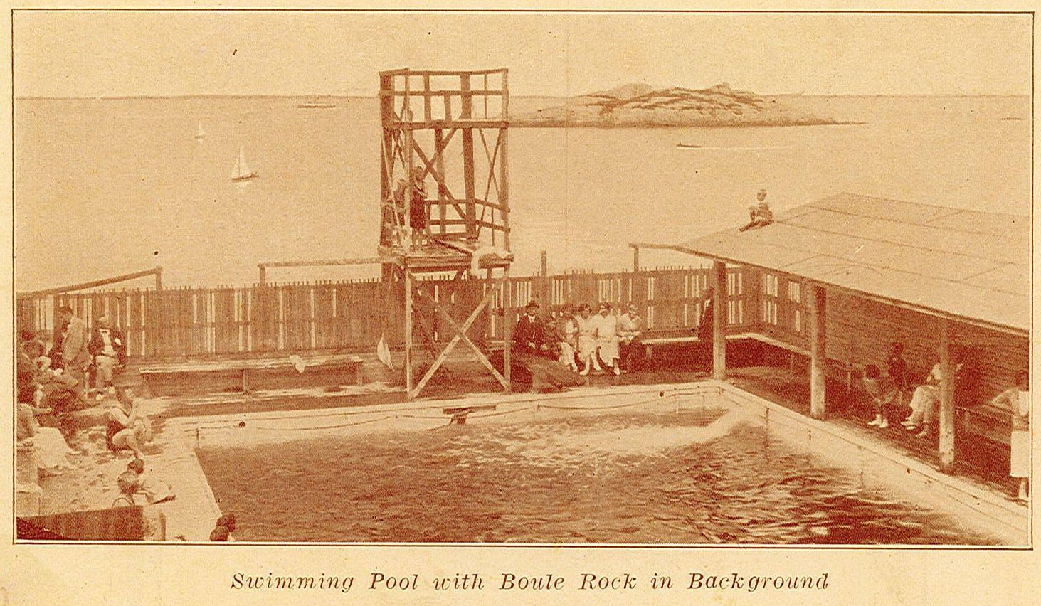 Many people sitting around a fenced pool with a large diving platform.