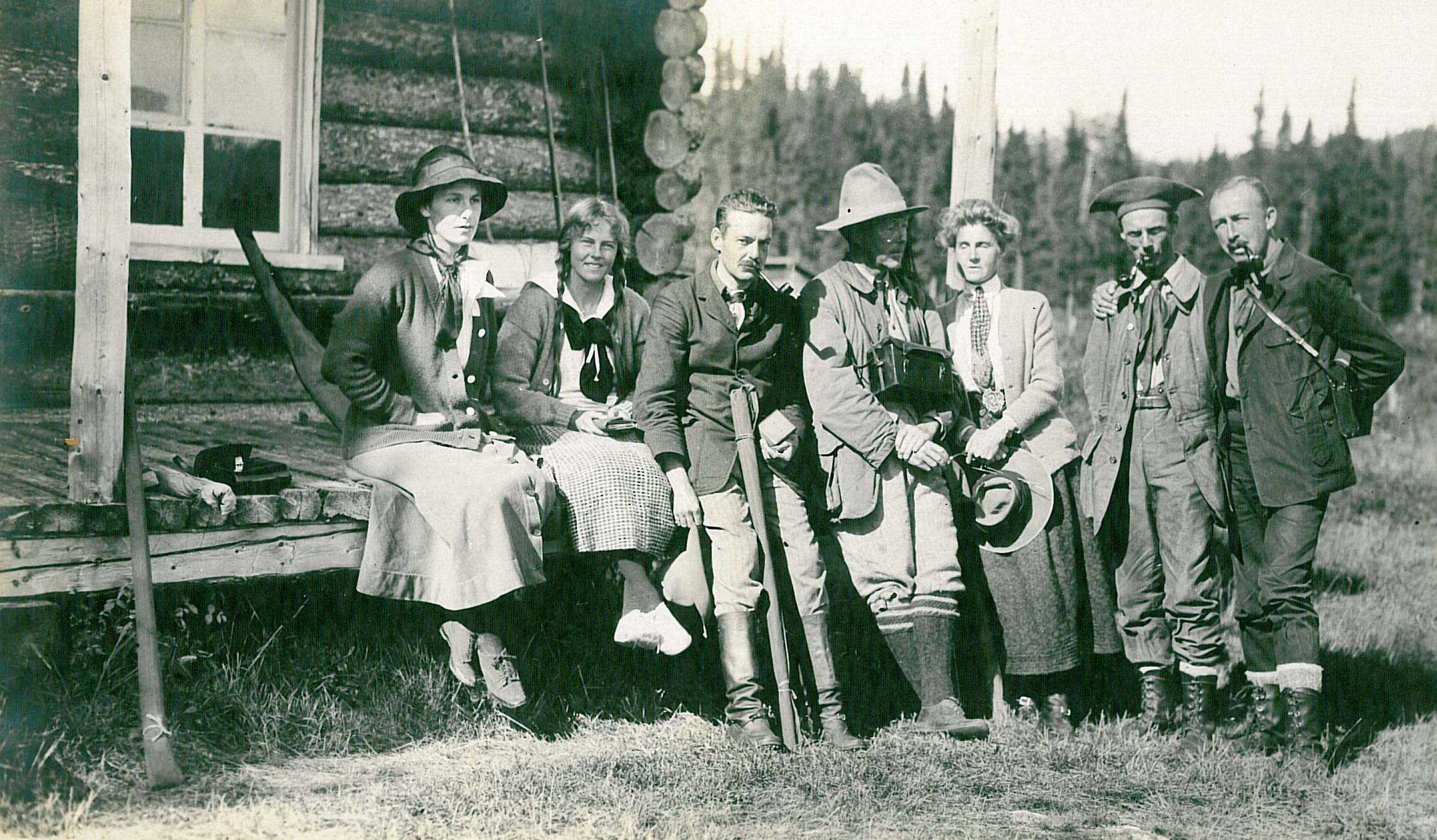 Four people are in front of a house with cedar shake walls, while two horses rest, each one attached to a four-wheeled carriage.