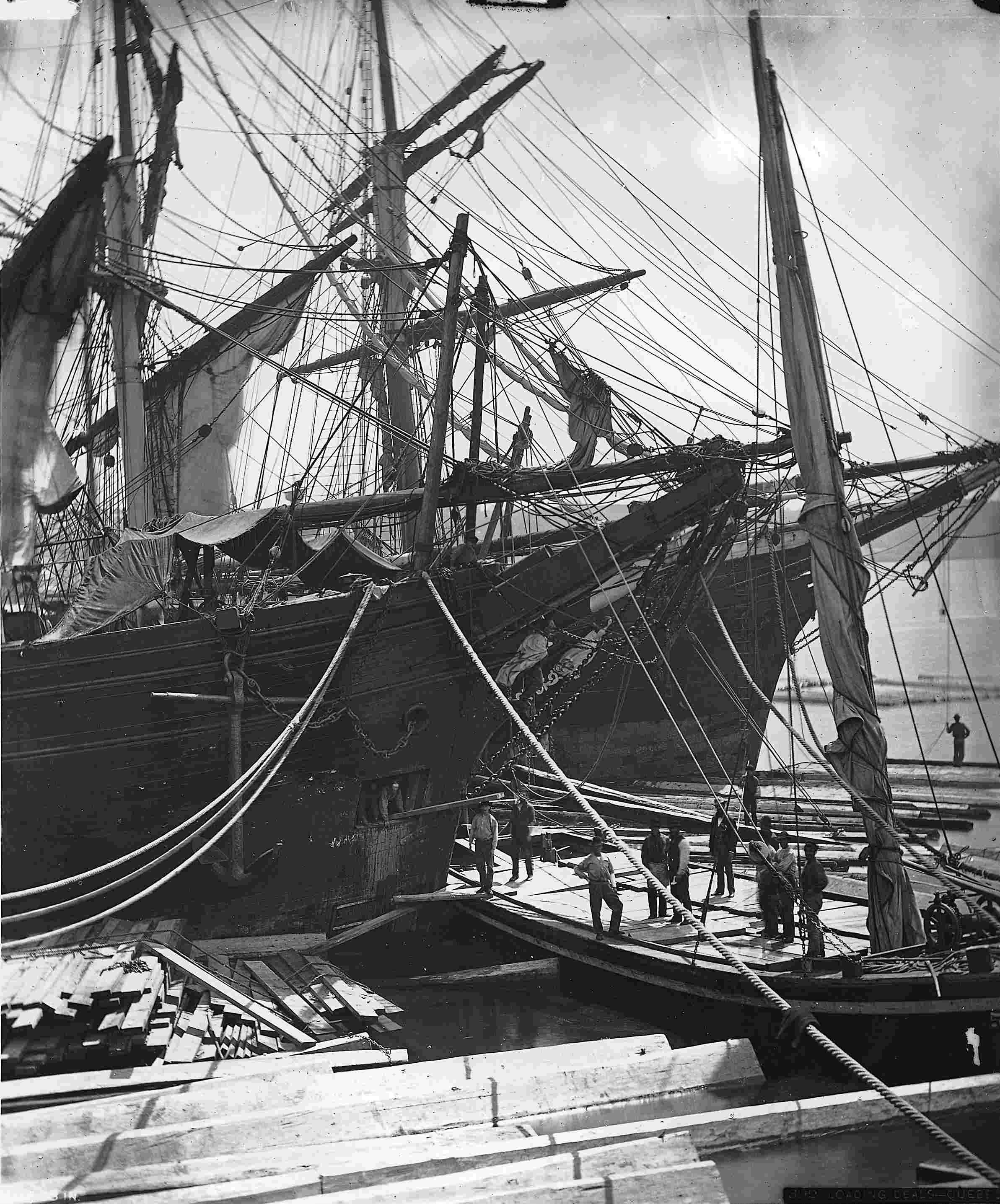 A dozen men unloading a large sailing vessel transporting lumber.