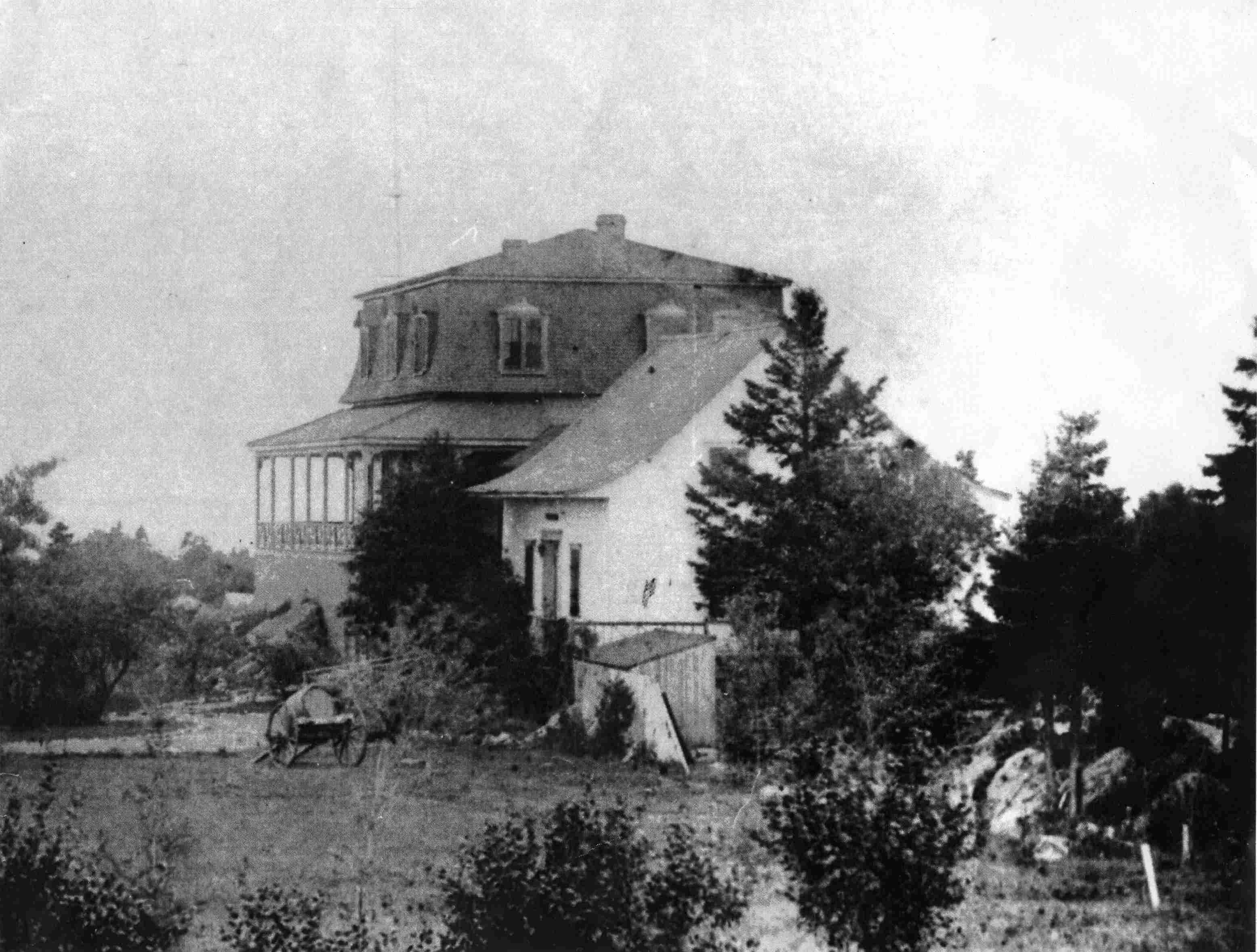 Photographie ancienne d’une maison de campagne entourée d’arbres, avec une falaise en arrière-plan.