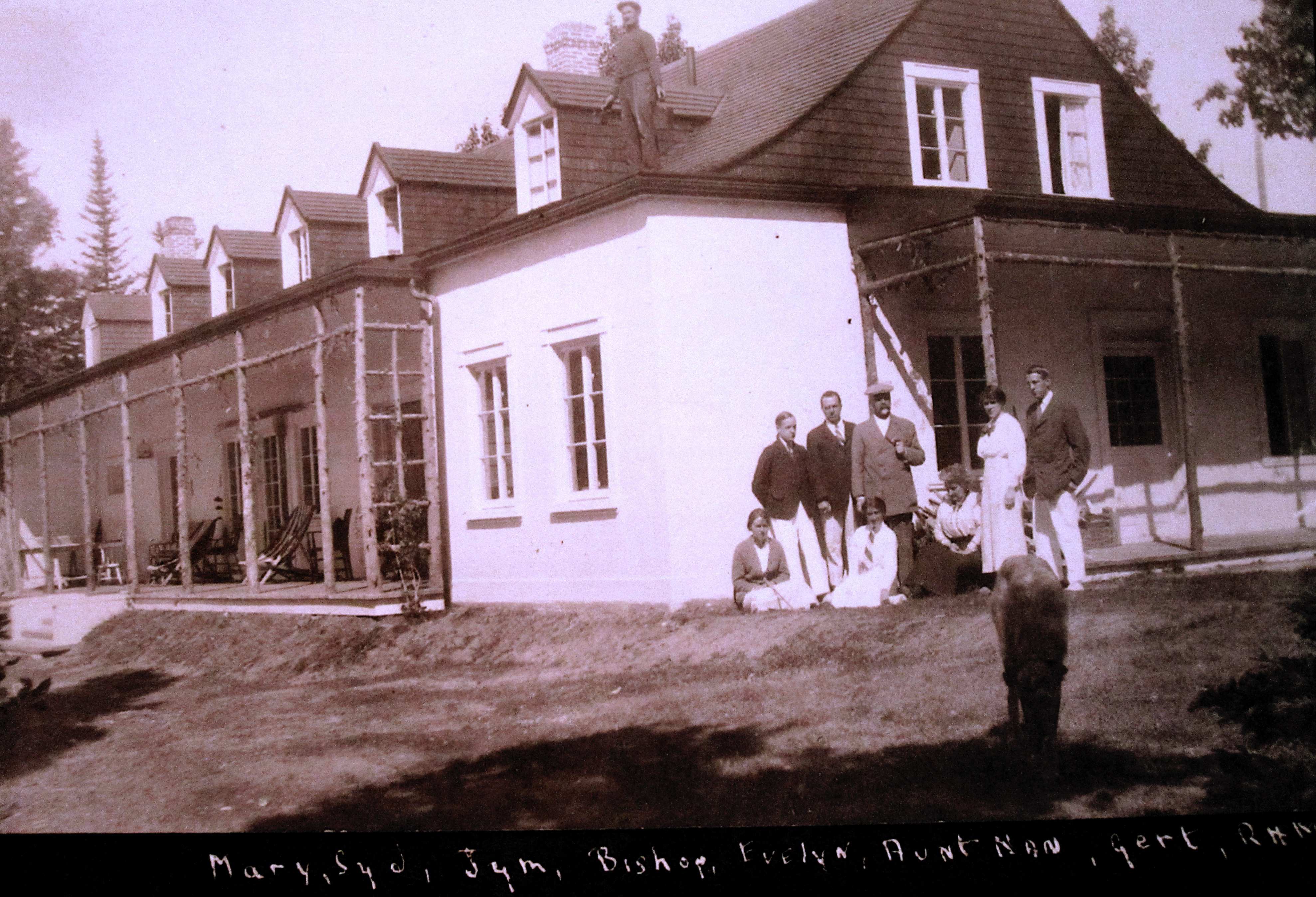 Several people pose in front of a large summer home; a workman is standing on the roof.