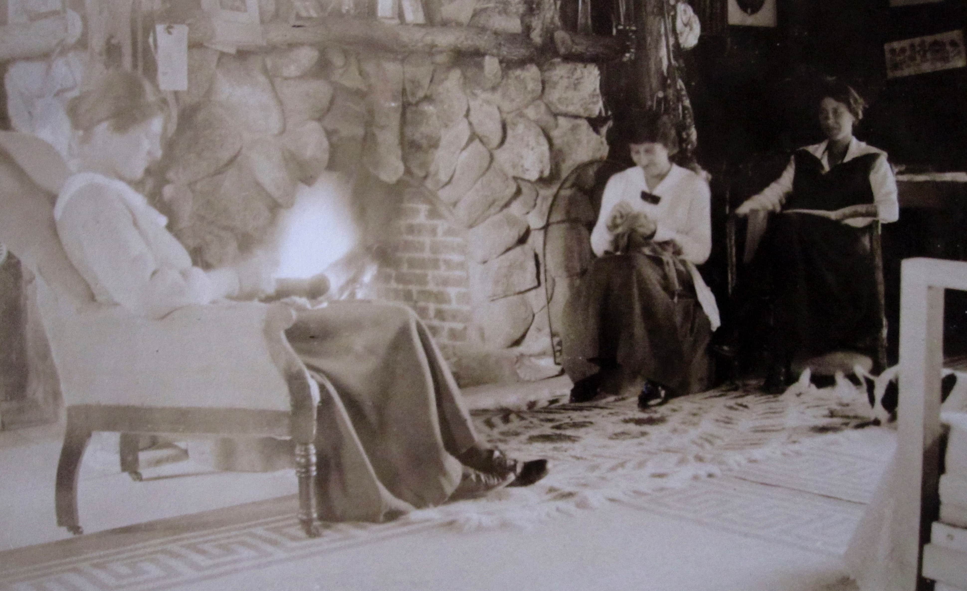 Three young women are reading and sewing in front of a fieldstone fireplace.