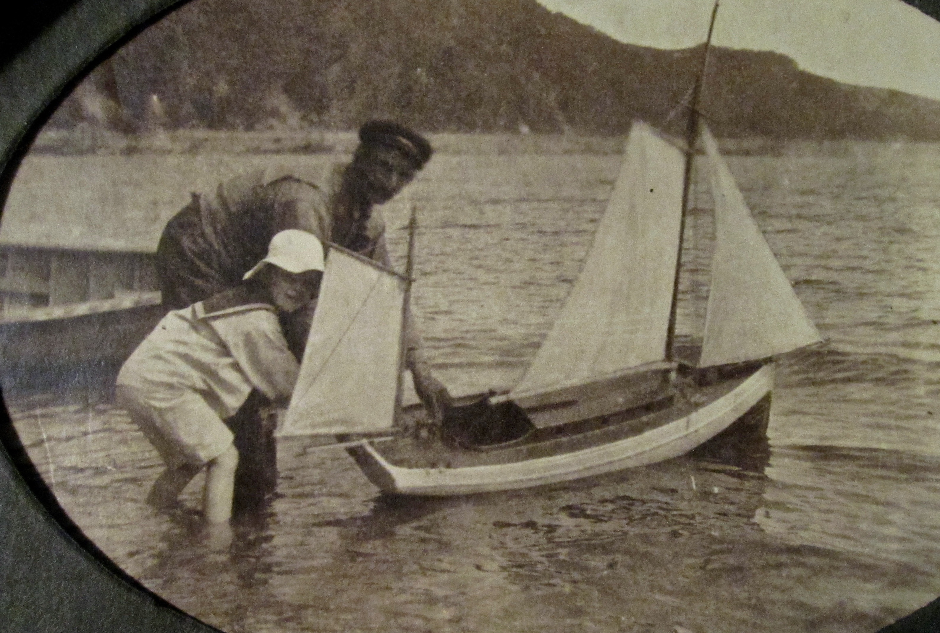 A man helps a boy in a sailor suit launch a model sailboat on the river.