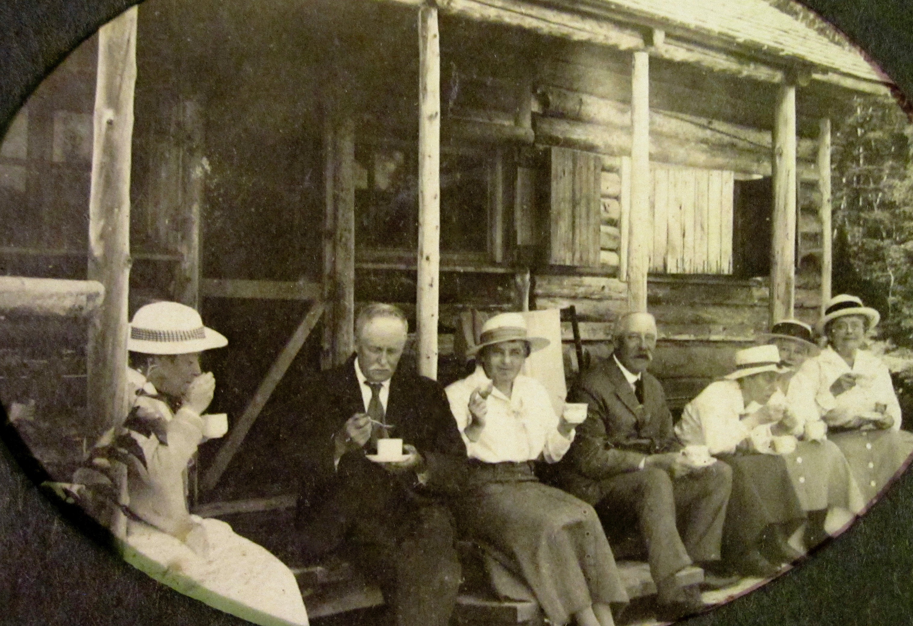 Seven middle-aged men and women drink tea on the porch of a rustic cabin.