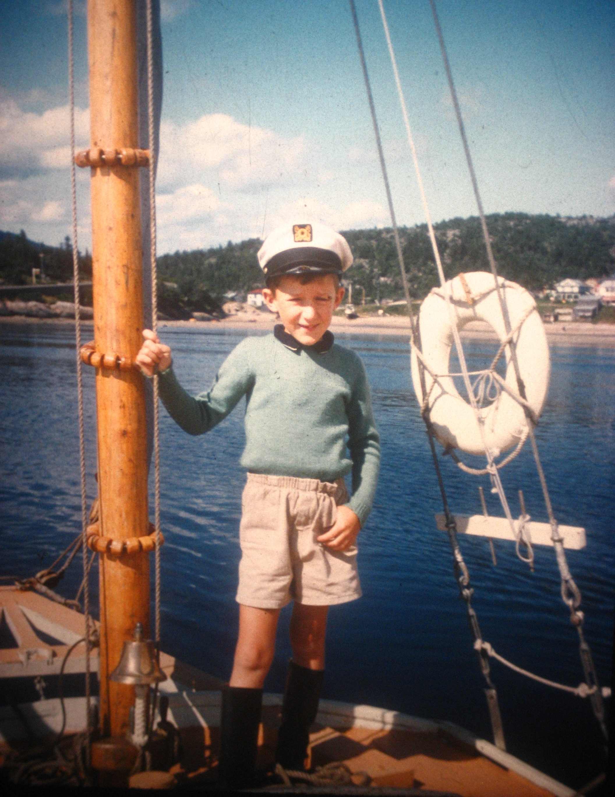 A colour photograph of a boy standing on the deck of a sailboat with the shore in the background.