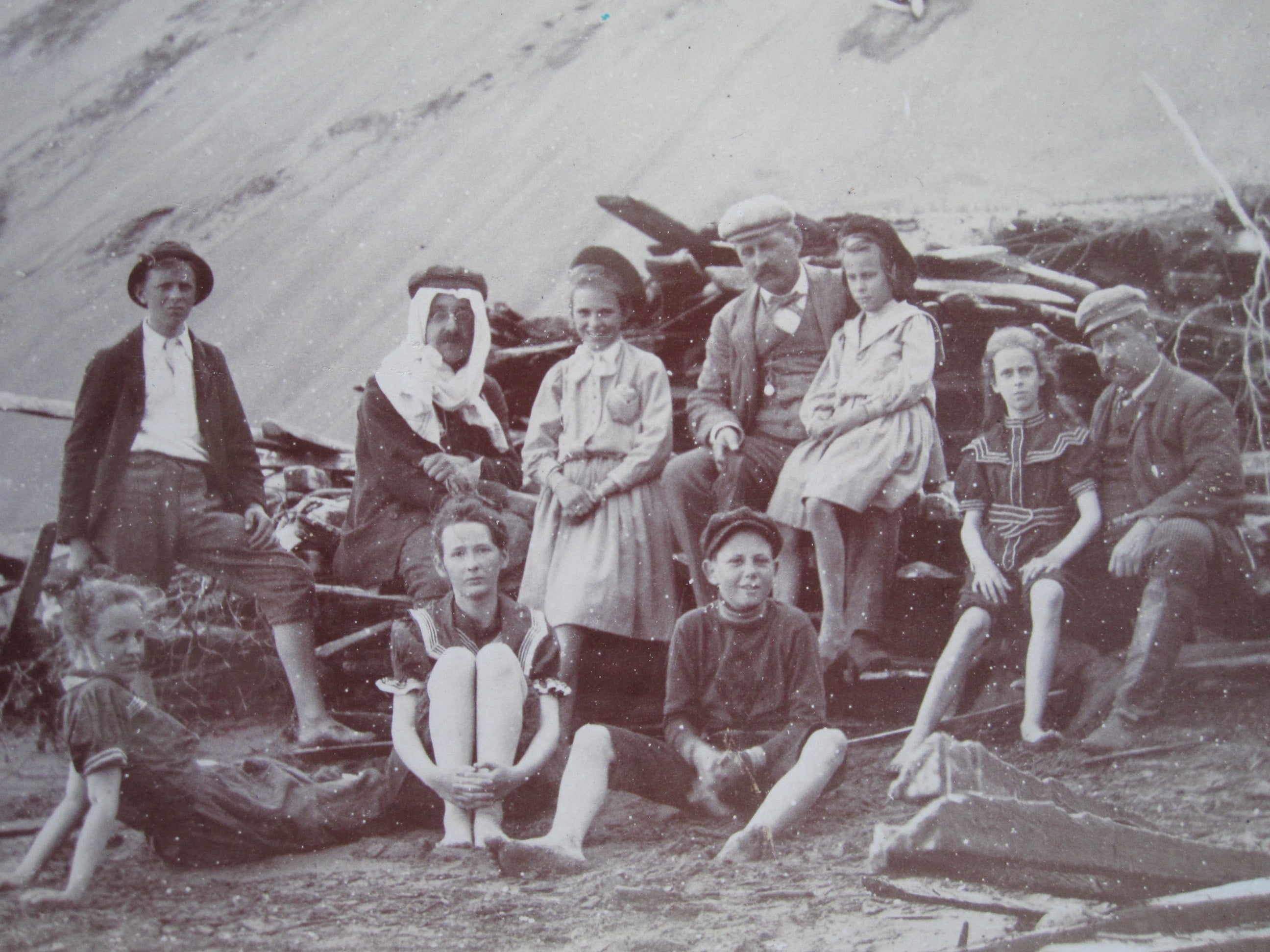 Ten people at the foot of a sand dune are sitting near a wood pile.