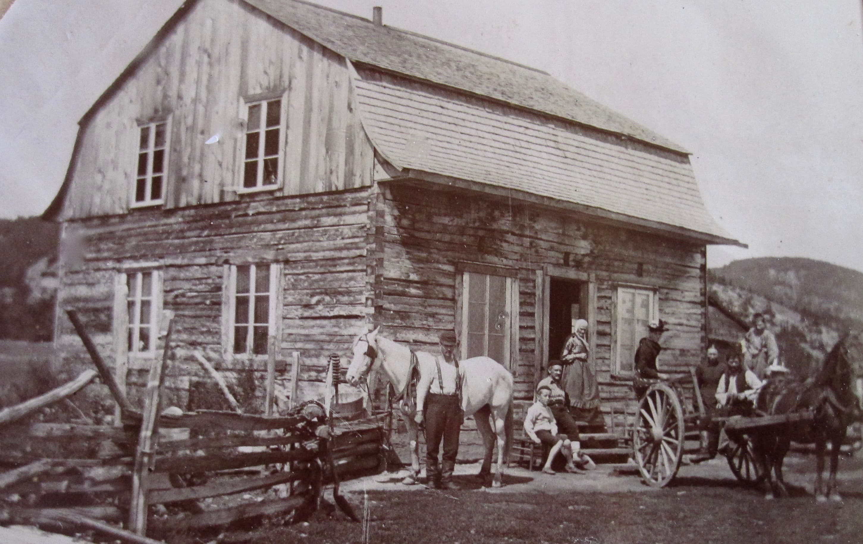 Quelques personnes posent devant une maison d'habitant construite en pièces sur pièces. Deux chevaux sont au repos.