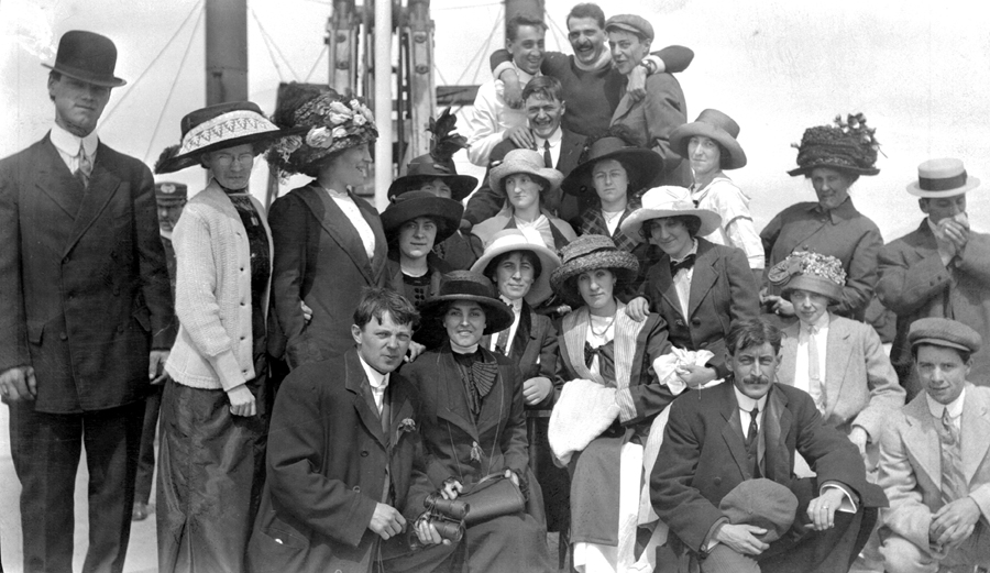 Un groupe d’hommes et de femmes; celles-ci portent des chapeaux à la décoration chargée.