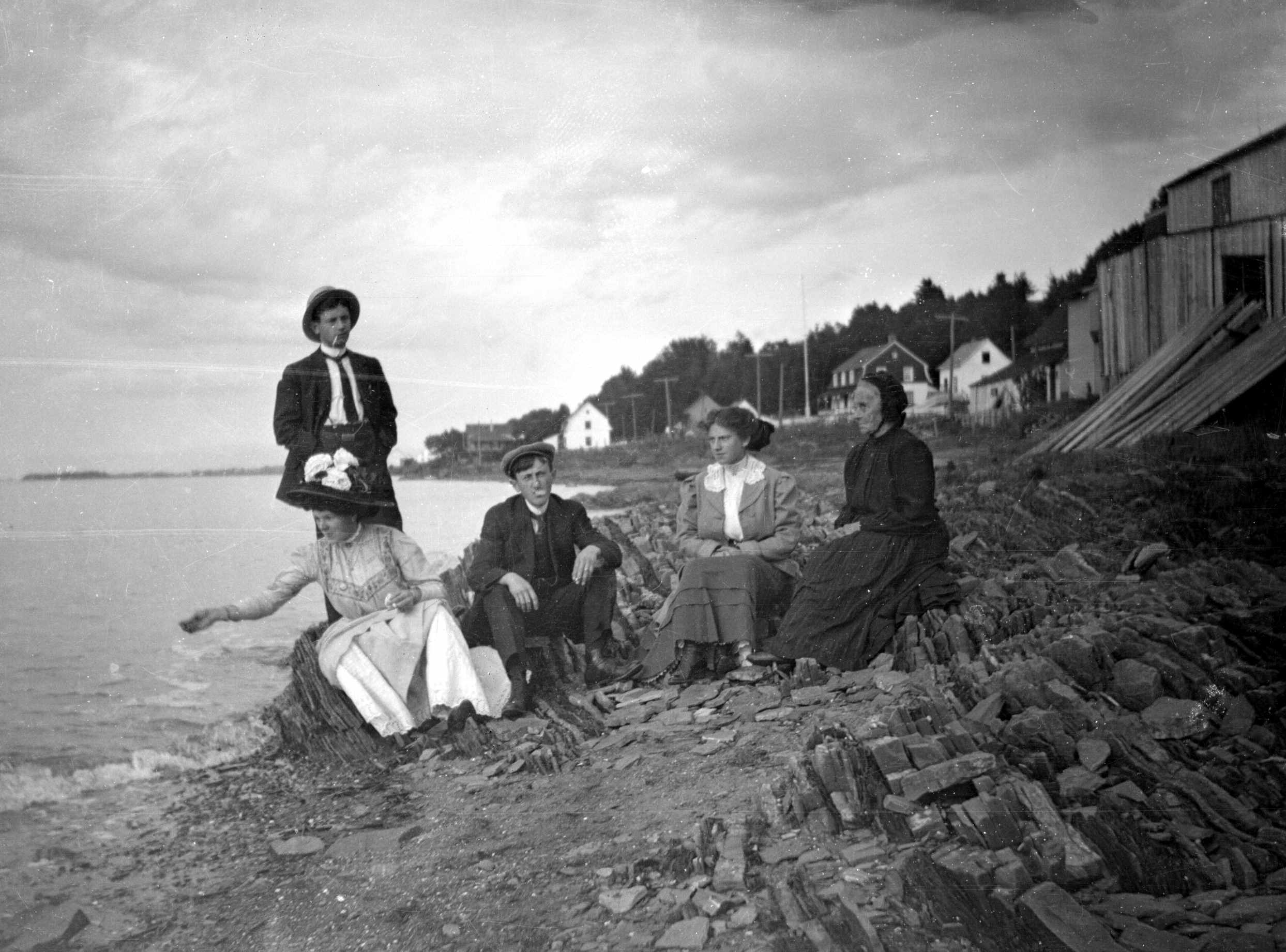 Adults, including an elderly lady, posing on a very rocky beach. One of the women is throwing bread to a bird out of view,