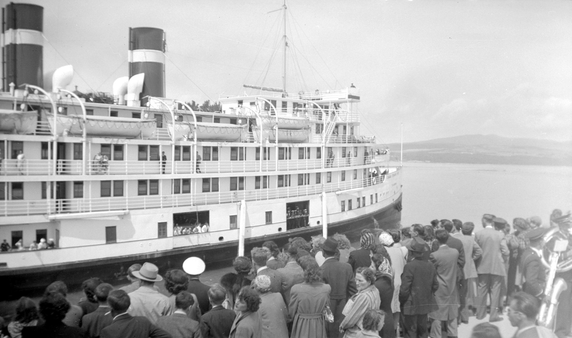 Dozens of passengers waiting on a wharf where a very large cruise steamer is moored.