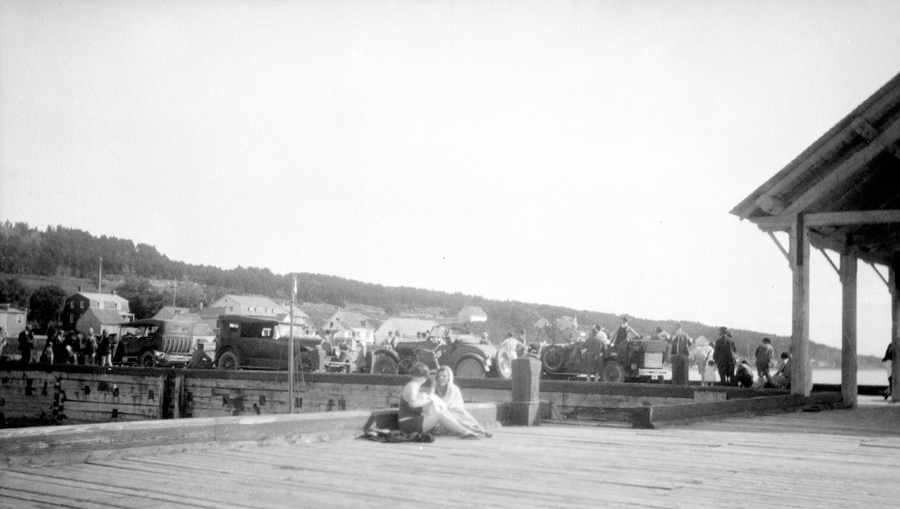 Two young women talking on an action-filled wharf, with several cars parked in the background.