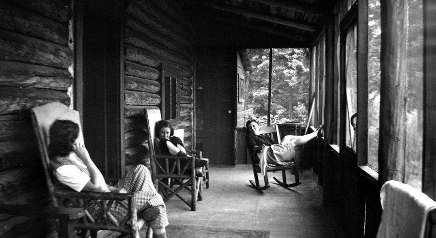 Three young women rocking on the porch of a log cabin.