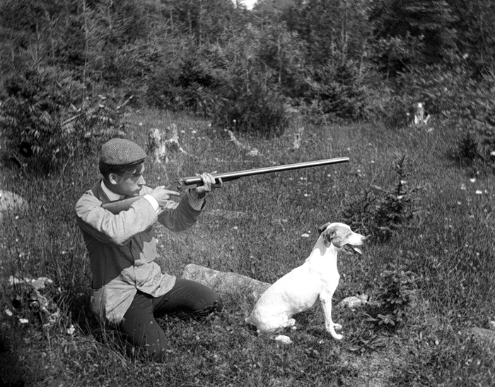 A young man is kneeling in the grass with his dog, hunting rifle in hand.