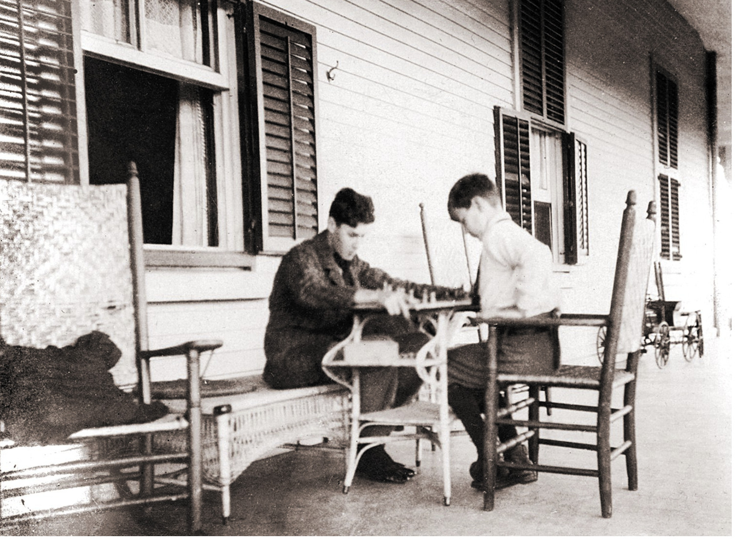 Two well-dressed teenage boys play chess on a veranda.