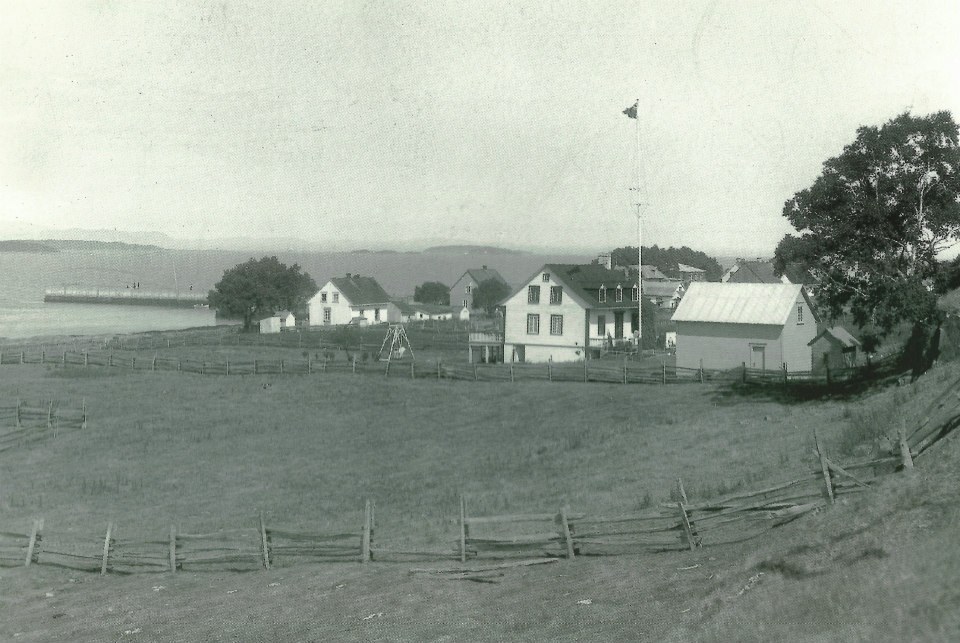 Beyond a field are a few houses and a wharf on the St. Lawrence River. The far shore is in the background.